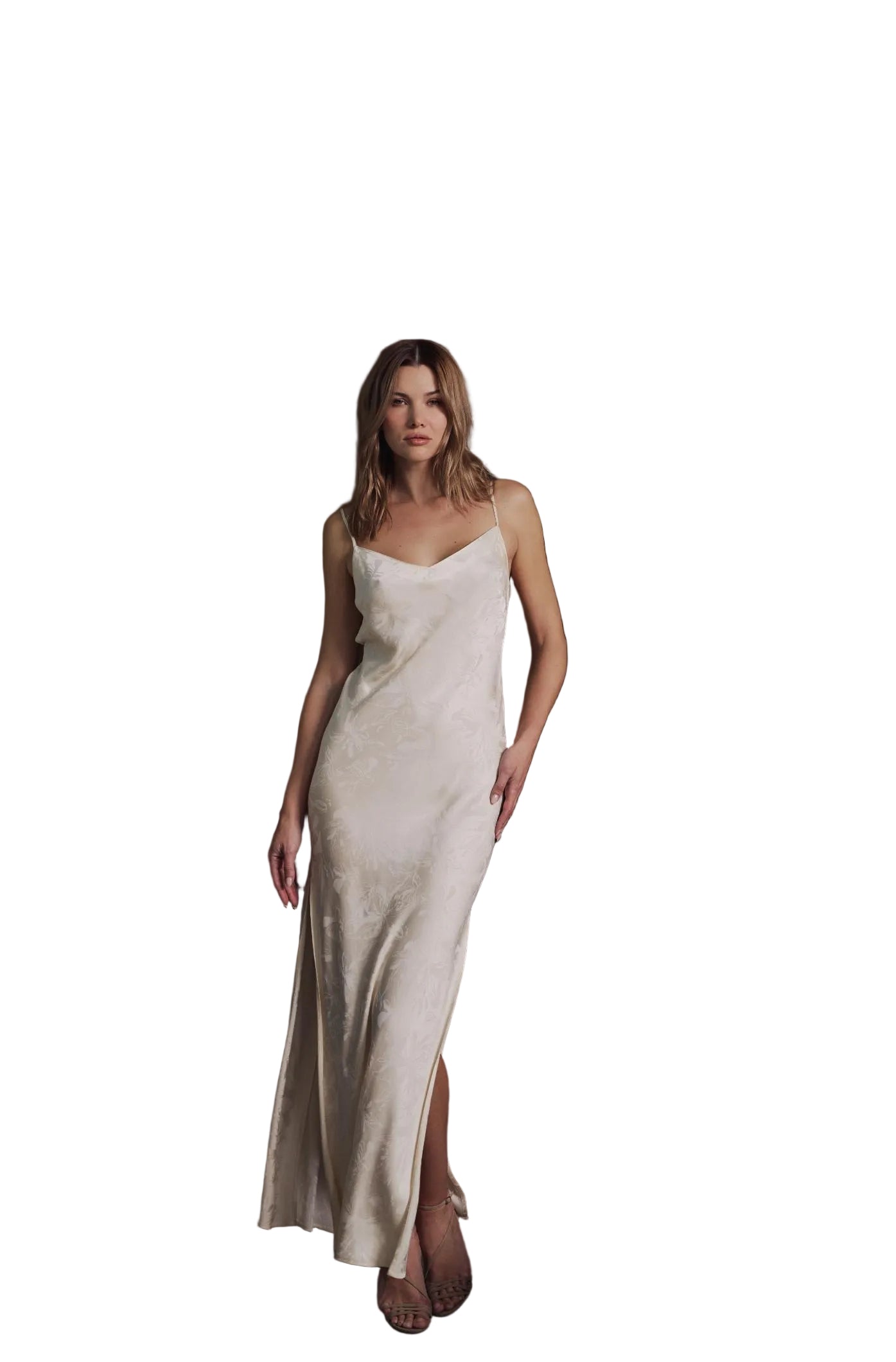 A woman wearing a sleeveless, floor-length champagne slip dress with a subtle jacquard pattern, standing in a modern interior with vertical wood slats and concrete stairs.