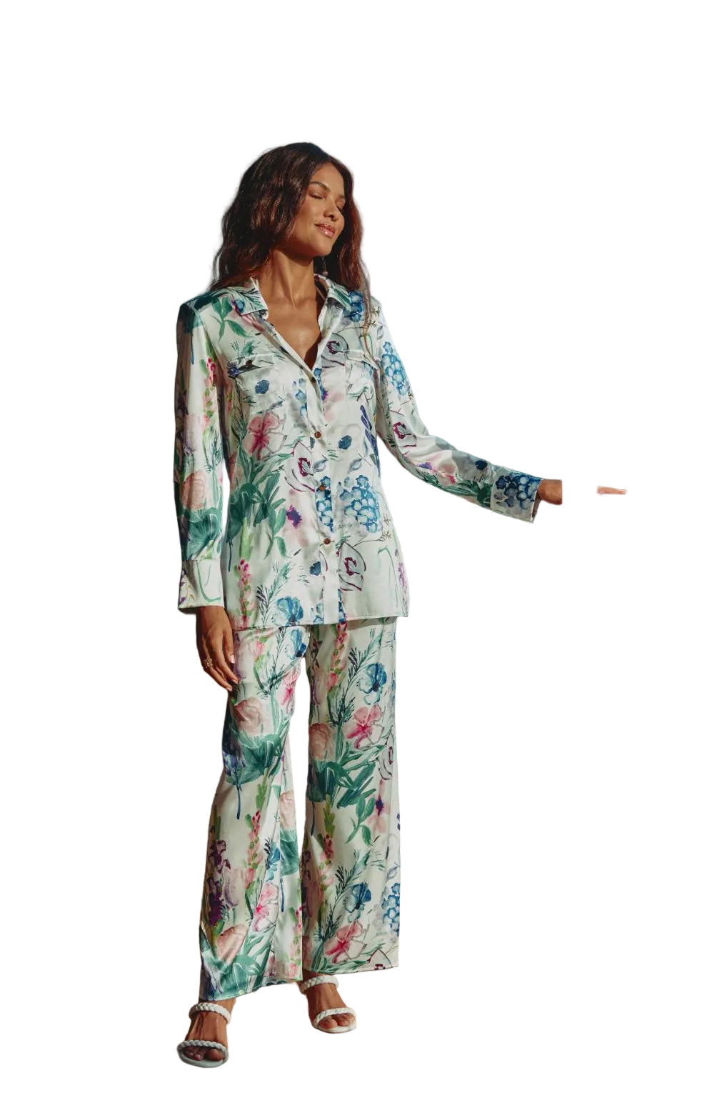 A woman stands on a sunlit balcony wearing a matching long-sleeve button-front shirt and wide-leg pants pajama set with a colorful floral print on a light background.