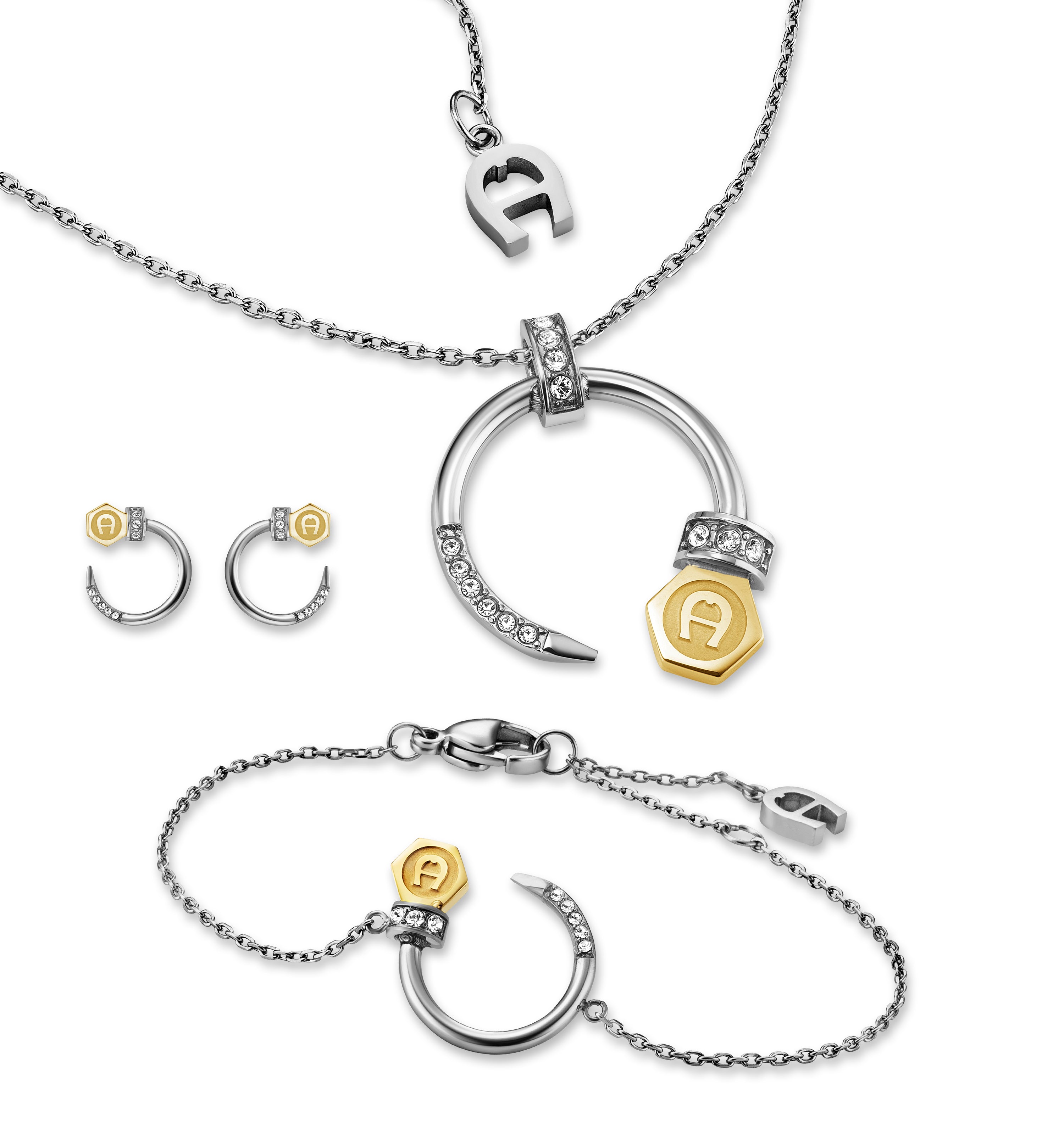 A studio display of the Aigner Matila jewelry set, showing the short cable chain necklace with its crystal-embellished crescent pendant, matching open-hoop earrings, and the adjustable chain bracelet featuring the two-tone gold hexagonal logo charm.