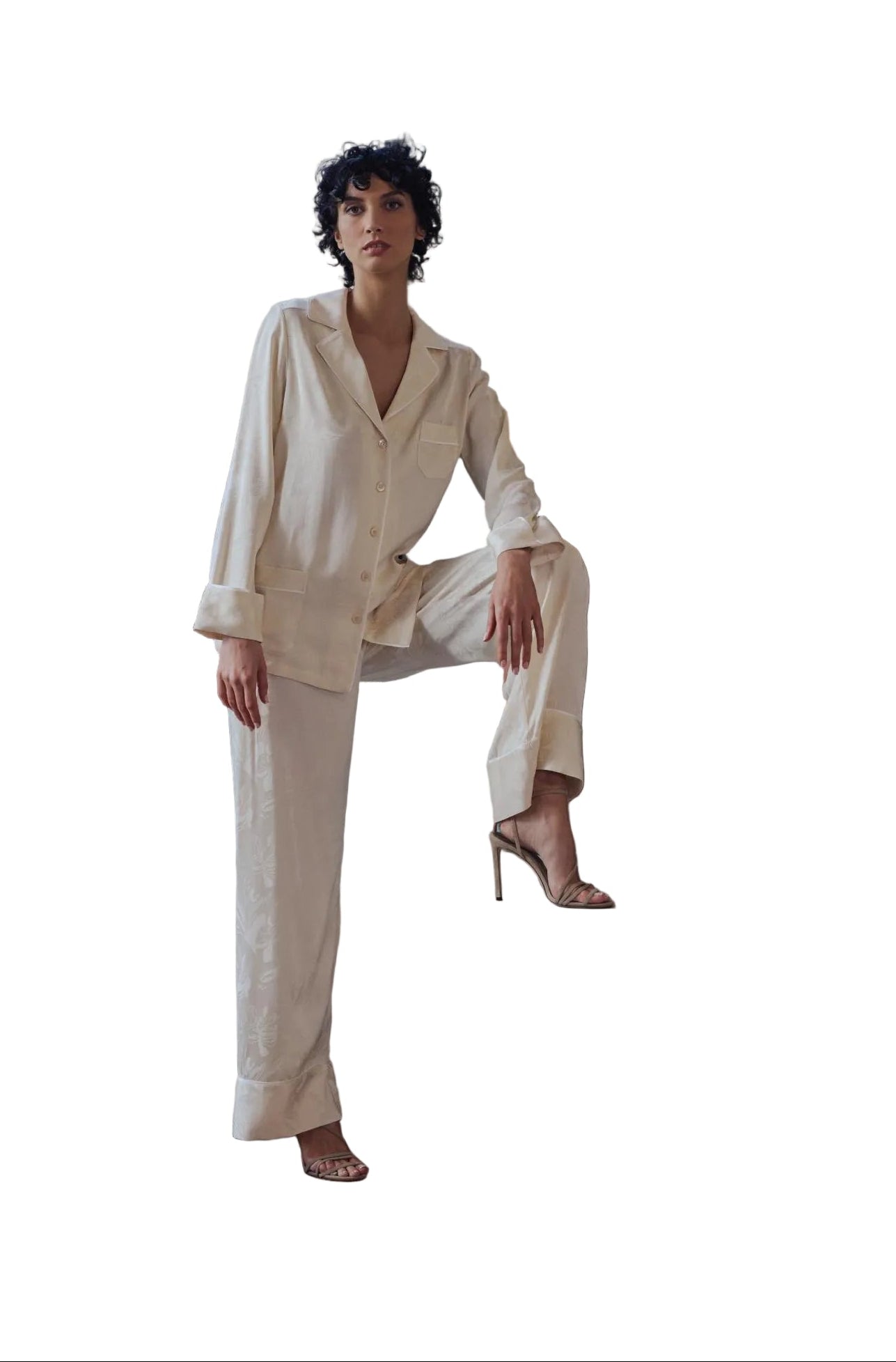 A woman models a matching cream-colored lounge set consisting of a button-down shirt and wide-leg trousers, posed on a concrete ledge in a minimalist modern room with a fireplace and large windows.