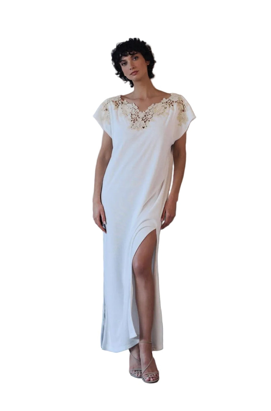 A woman stands in front of a large concrete fireplace wearing a long white slip dress with gold embroidery on the bodice and a high slit.