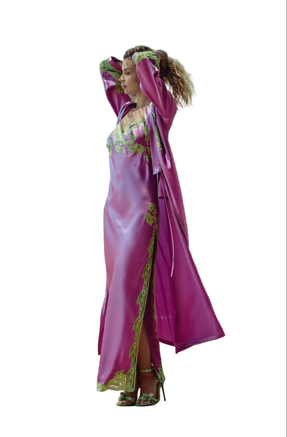 A woman stands on a balcony wearing a flowing magenta-purple robe with green embroidered trim and a matching headpiece, with a mountain and greenery in the background.