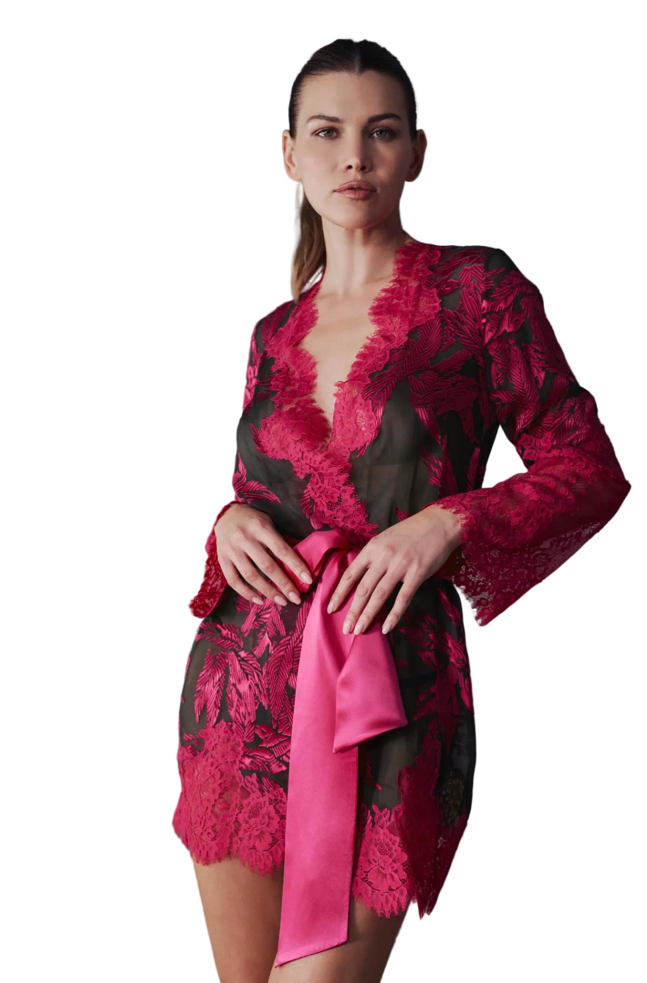 A woman models a short, long-sleeved lace robe in vibrant magenta pink and black floral lace, featuring a matching satin tie belt, posed in front of a gray wall and light gray sofa.
