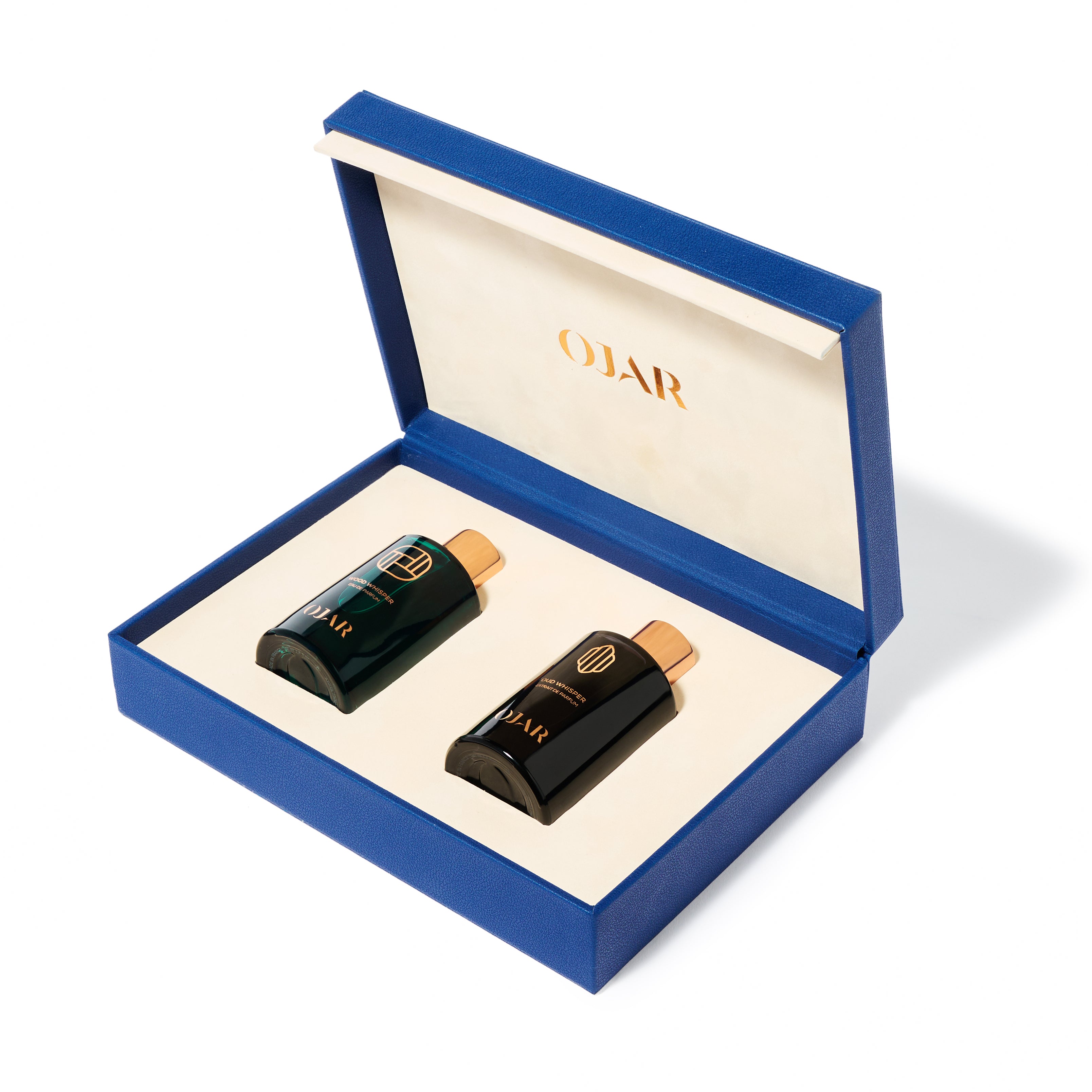 An open OJAR "The Whisper Set" gift box featuring a vibrant cobalt blue exterior and a cream-colored velvet interior. The box holds two 15ml fragrance bottles with copper caps: one in translucent emerald green (Wood Whisper) and one in opaque midnight black (Oud Whisper). The brand logo "OJAR" is printed in gold on the inside of the lid.