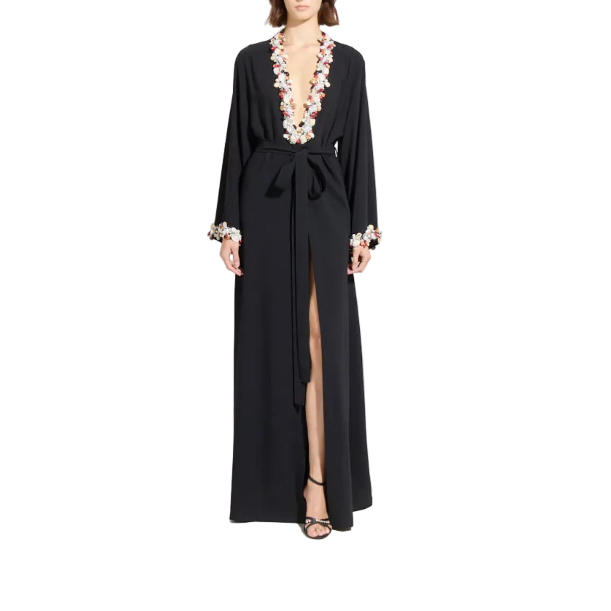 A woman modeling a long black wrap-style maxi dress with a deep V-neckline, high front slit, and white floral embroidery around the neckline and cuffs, shown against a plain white background.