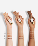 Three hands of different skin tones holding Guerlain Terracotta Joli Teint tubes next to arm swatches labeled from shades 10 to 50, available at blue salon.