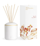 White cylindrical reed diffuser with a gold-toned band, containing natural wooden reeds, next to an illustrated white box.