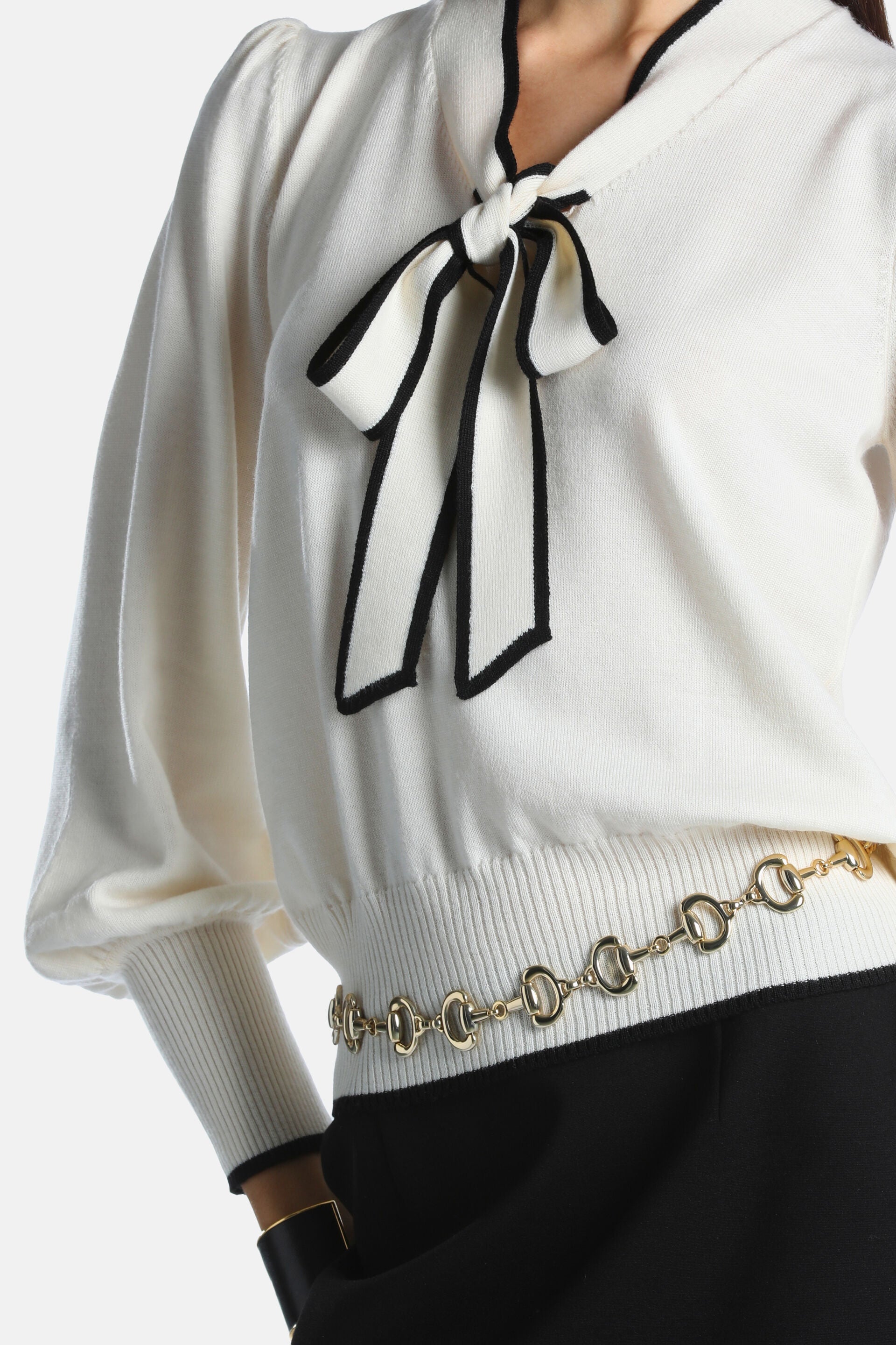 Cream knit pullover with black trim. Features a V-neckline with a tied bow, puffed sleeves, ribbed cuffs, and a ribbed hem.