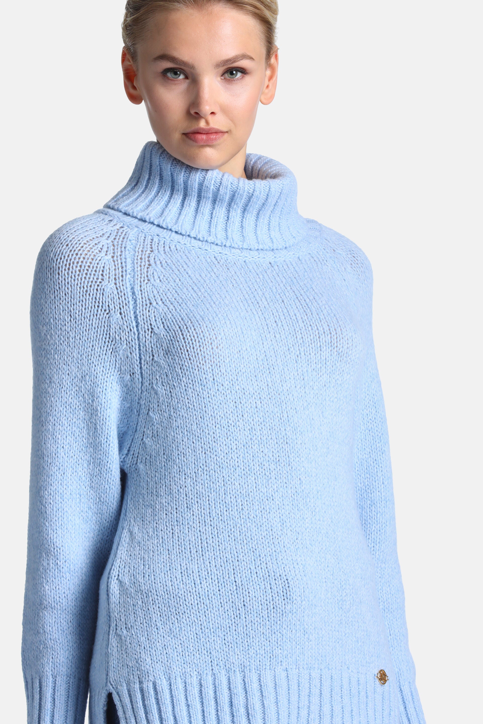 Light blue chunky knit turtleneck sweater with ribbed collar, raglan sleeves, ribbed cuffs, side slits, and a gold-toned embl