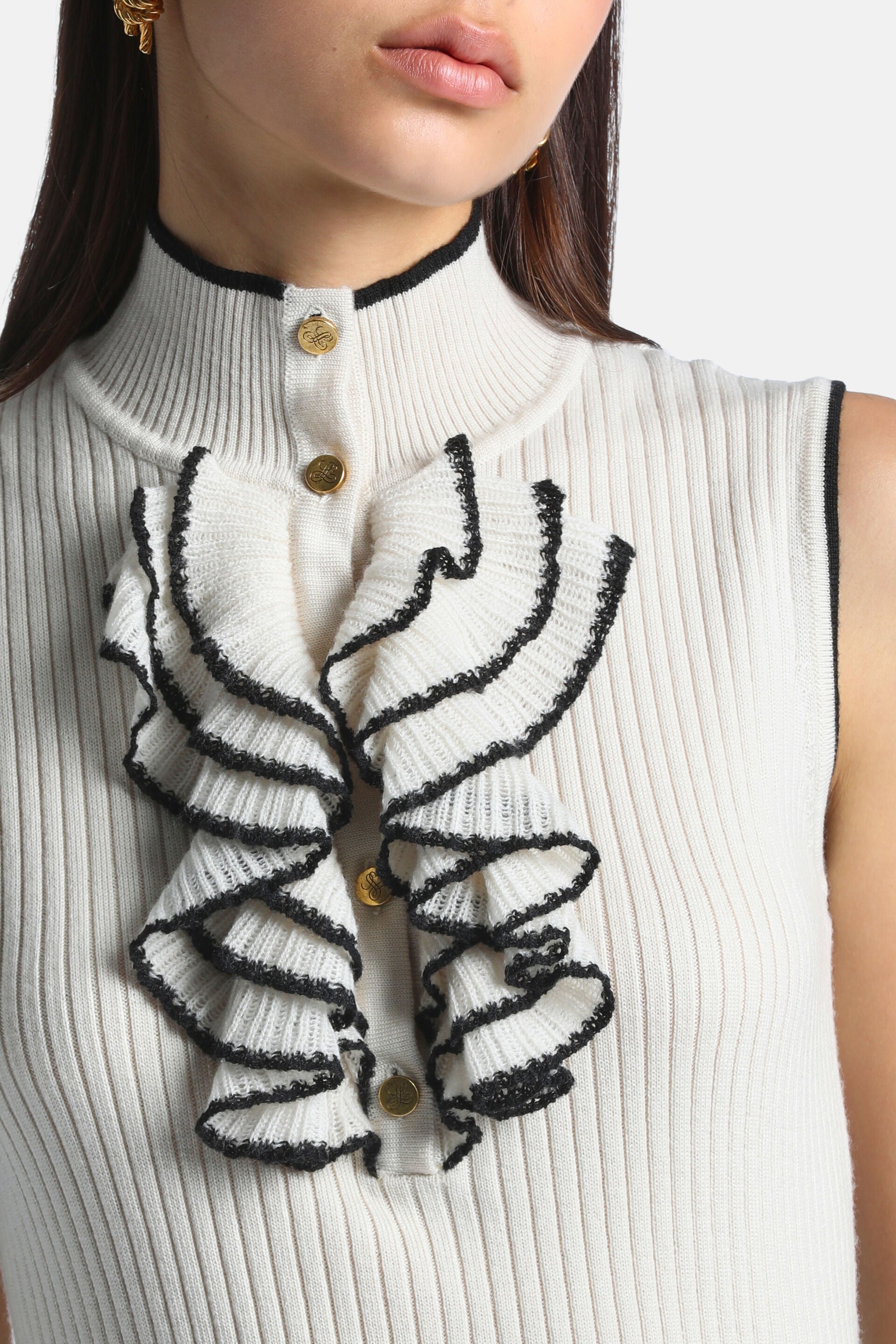 Cream ribbed knit sleeveless pullover with black trim. Front has gold-toned buttons and cream ruffles with black edges.