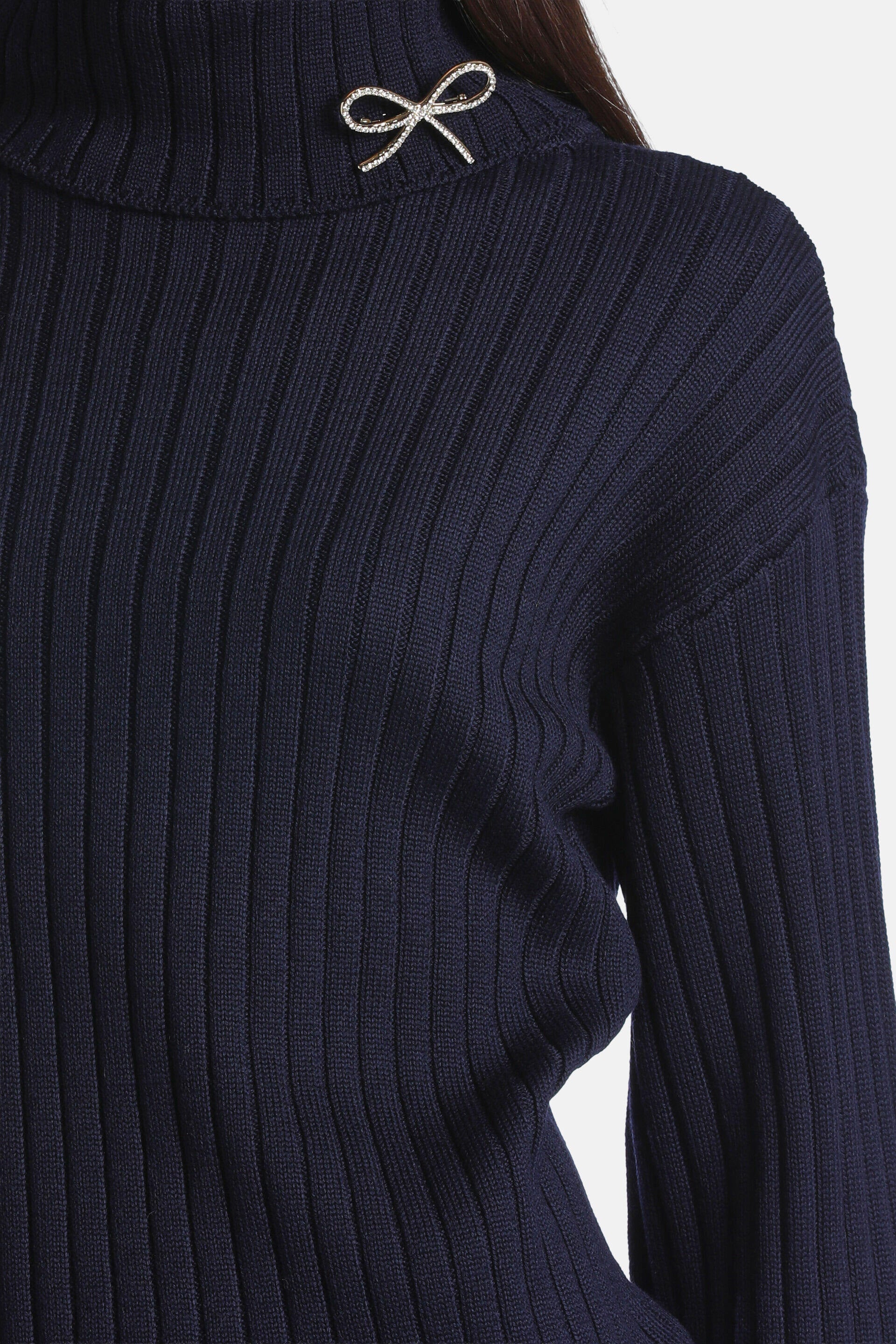 Navy blue ribbed knit turtleneck pullover with a silver-toned, rhinestone-studded bow brooch on the left side of the neck.