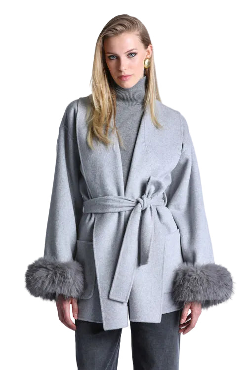 A woman modeling a light gray belted wrap coat with gray faux-fur trimmed cuffs, worn over a gray turtleneck sweater and dark gray pants, against a plain light gray background.