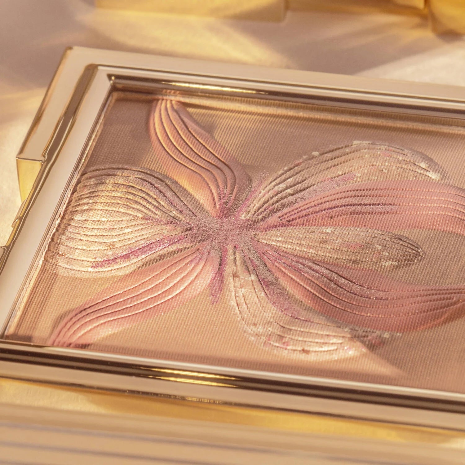 Gold-toned rectangular makeup compact with shimmery bronze powder featuring a raised pinkish-bronze orchid design.