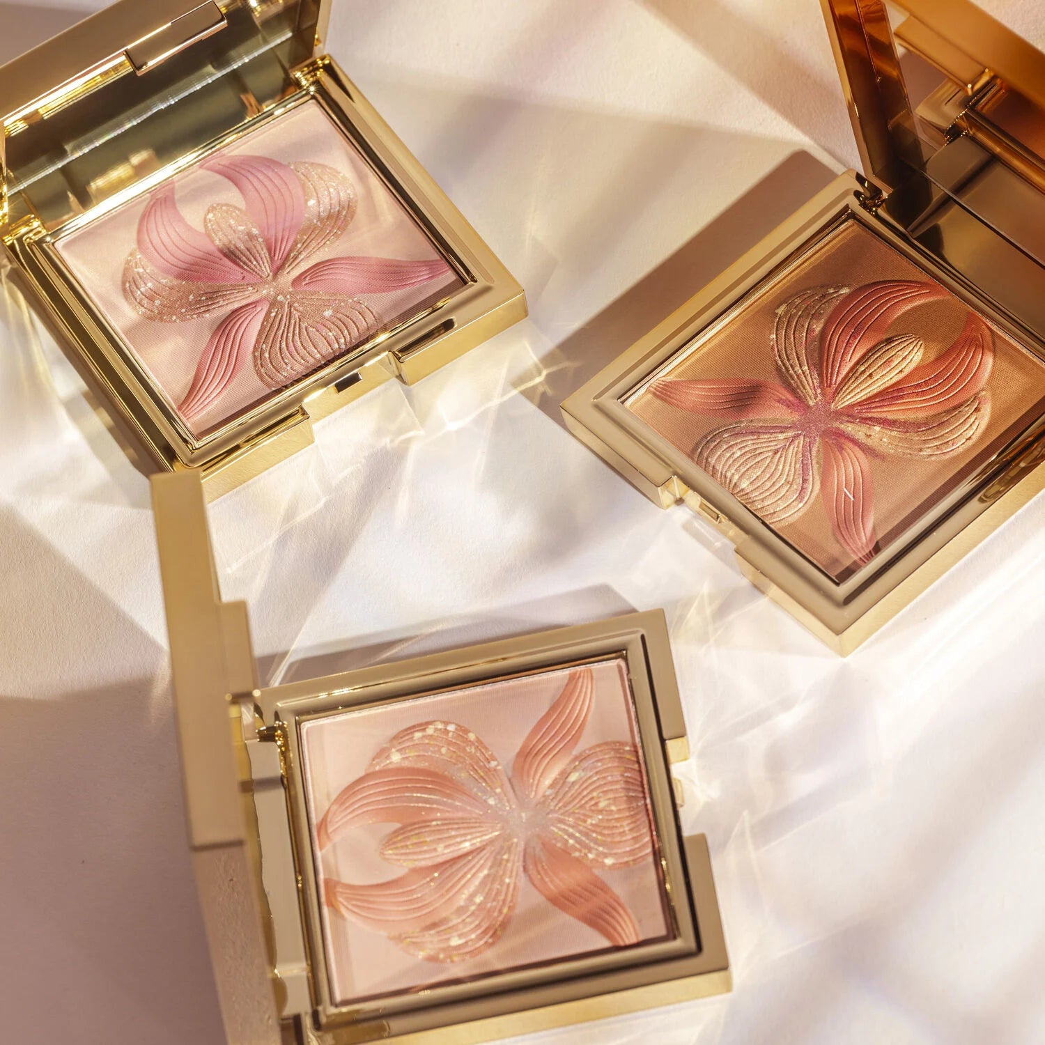 Square gold-toned compact with bronze pressed powder featuring a raised orchid design with pink and gold-toned glitter accent
