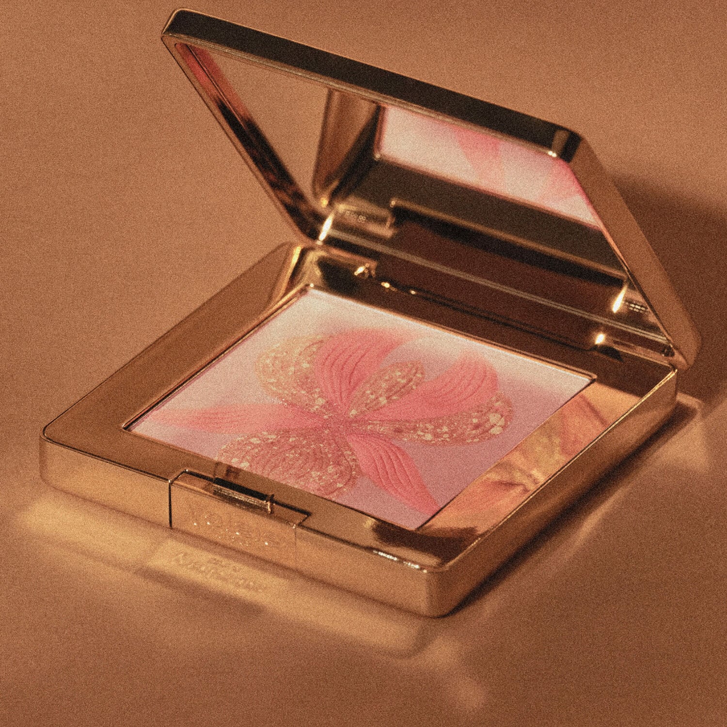 Gold-toned compact, open to reveal a mirror and pressed powder. Powder has a peachy-pink flower with glittery gold-toned deta