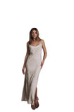 A woman wearing a sleeveless, floor-length champagne slip dress with a subtle jacquard pattern, standing in a modern interior with vertical wood slats and concrete stairs.