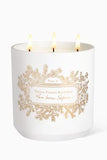 White cylindrical candle with three lit wicks, featuring a gold-toned pine branch label design on the front.