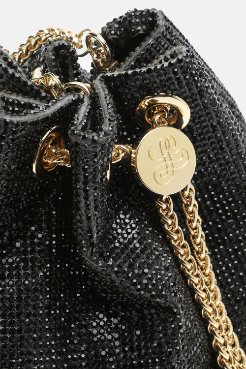 Black drawstring bag covered in black rhinestones, with gold-toned chain straps and a round gold-toned logo medallion.