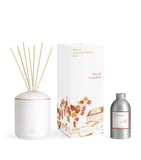 White ceramic diffuser with gold-toned band, wooden sticks, a white box with red/gold botanical art, and a silver-toned refil