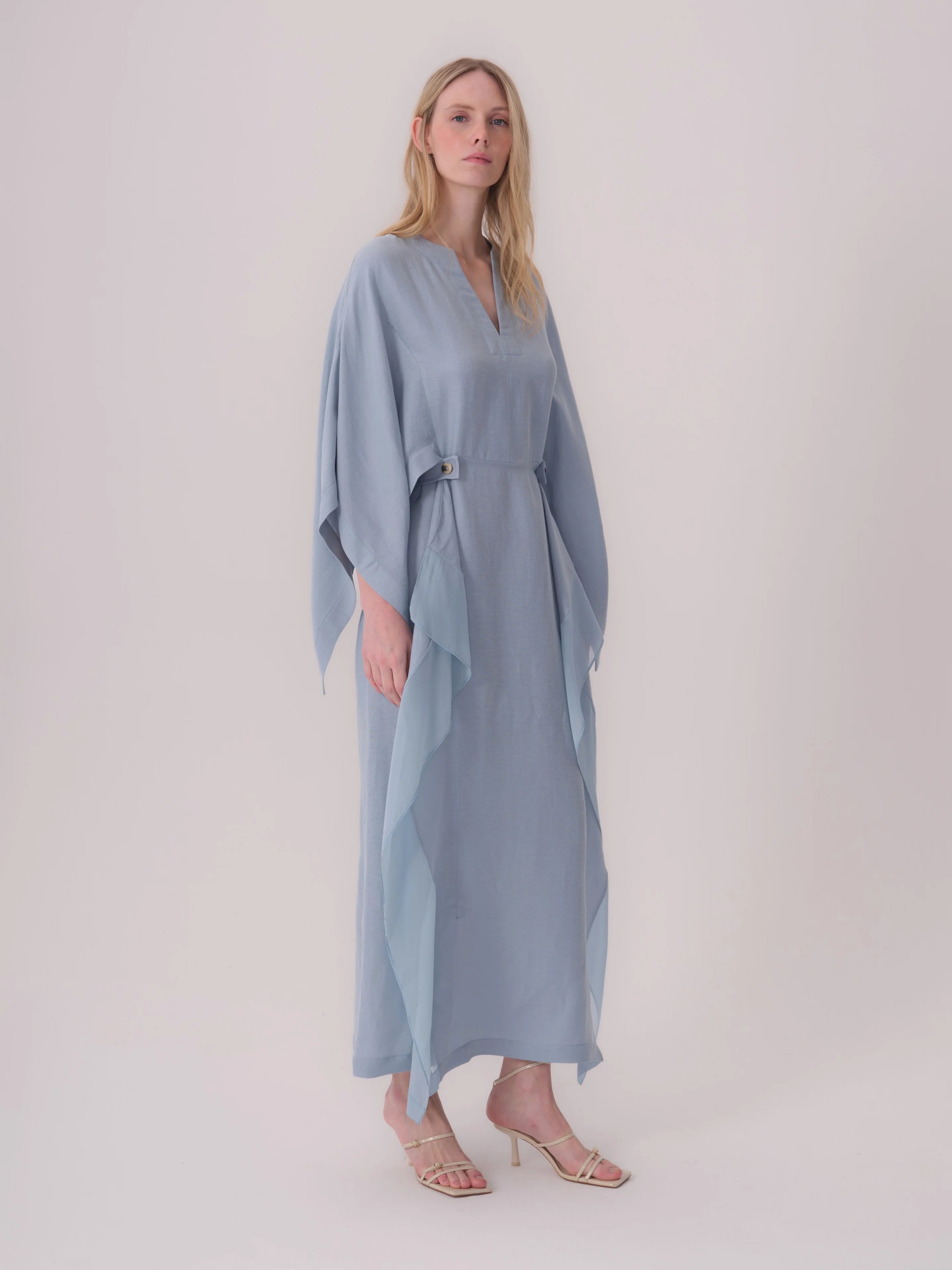 Light blue maxi dress with V-neck, wide flowing sleeves, a gold-toned buckle at the cinched waist, and sheer lighter blue sid