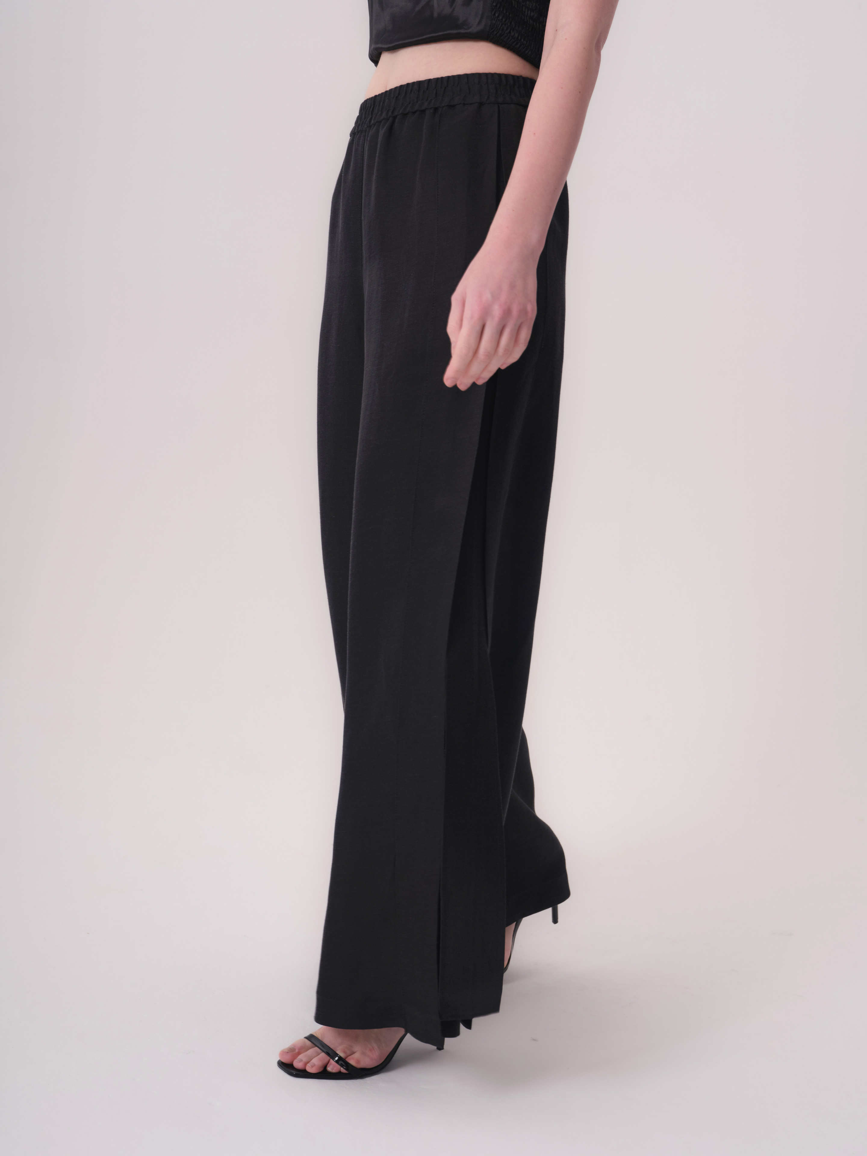 Black wide-leg trousers with an elasticated waist and a subtle sheen. The left leg has a short slit at the ankle.