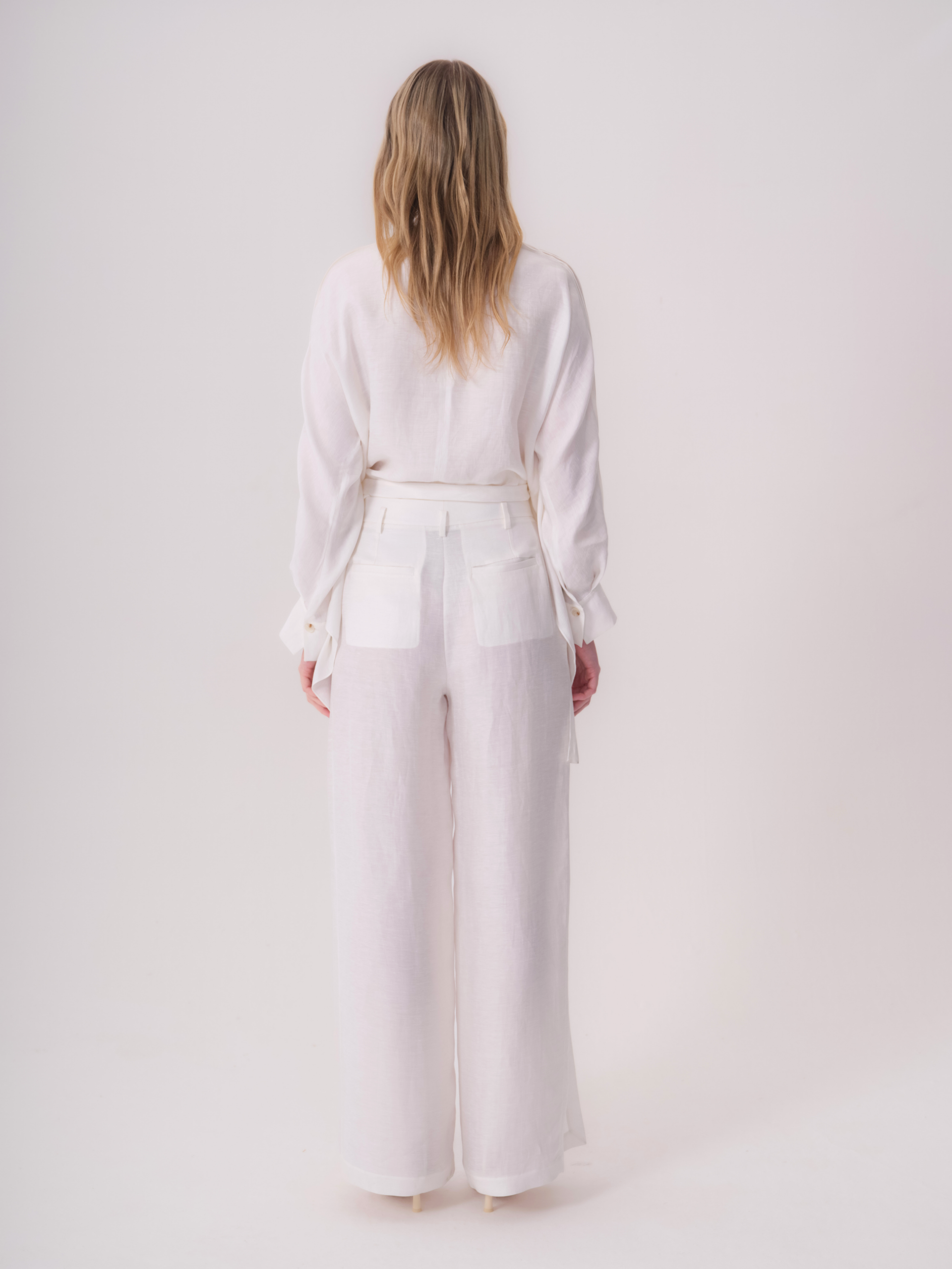White wide-leg linen trousers with a high, wide waistband and two back patch pockets. Ties hang from the sides of the waistba