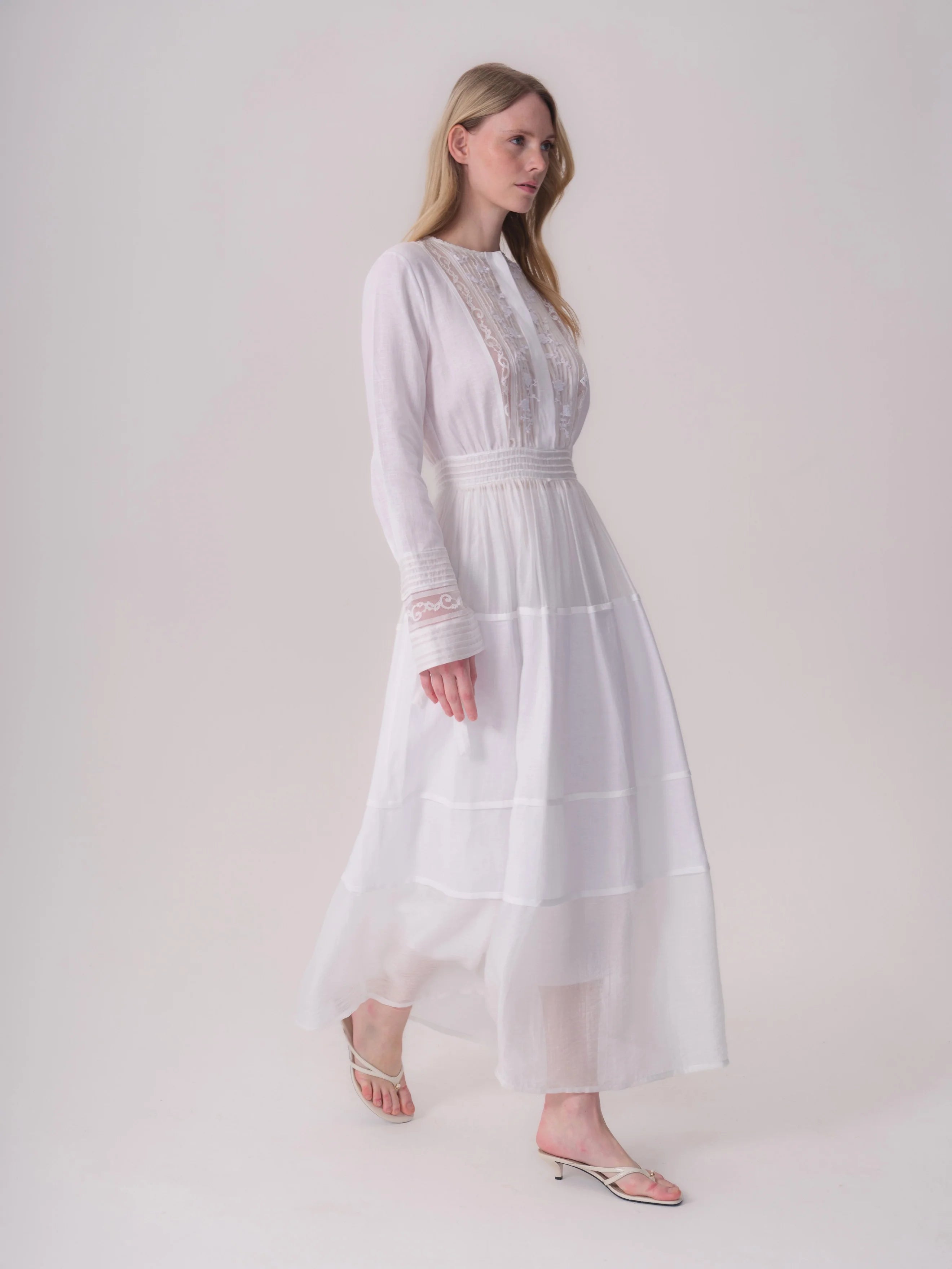 White long-sleeve maxi dress with a band collar, lace paneling on the bodice, shirred waist, and tiered skirt.