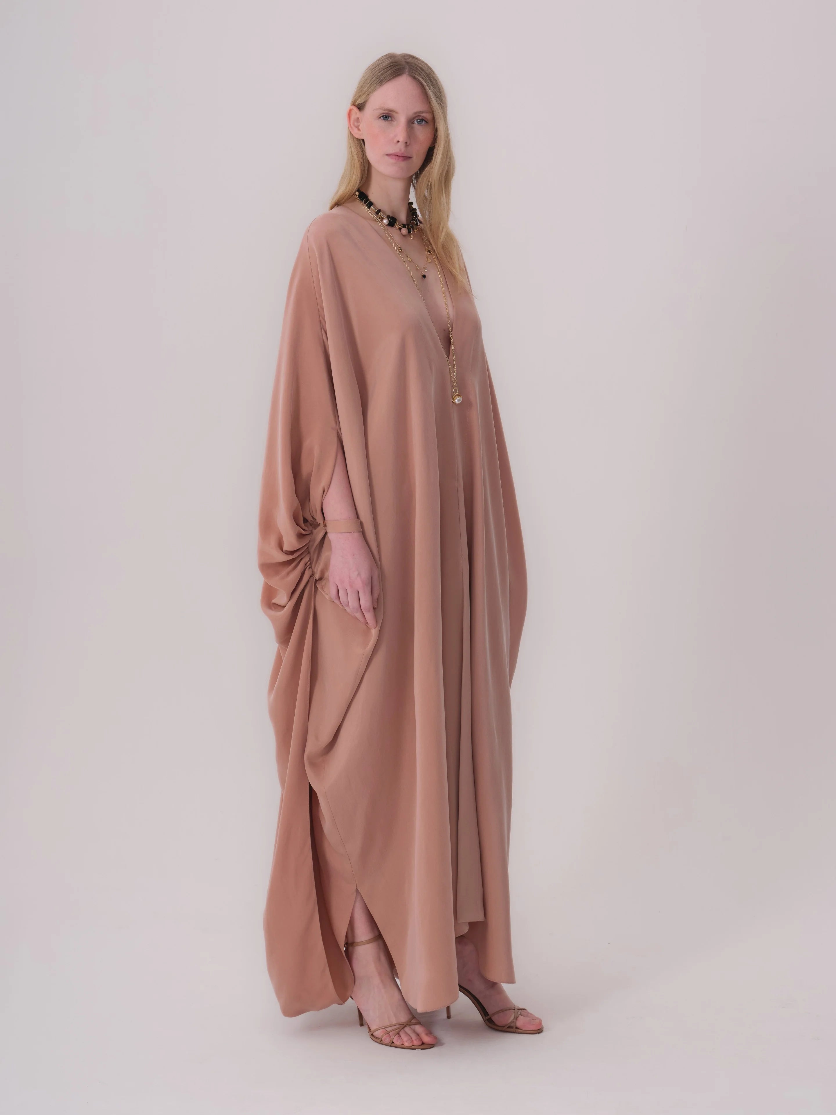 Dark beige flowing abaya with V-neck, wide dolman sleeves gathered at wrists, draped left side, and side leg slits.