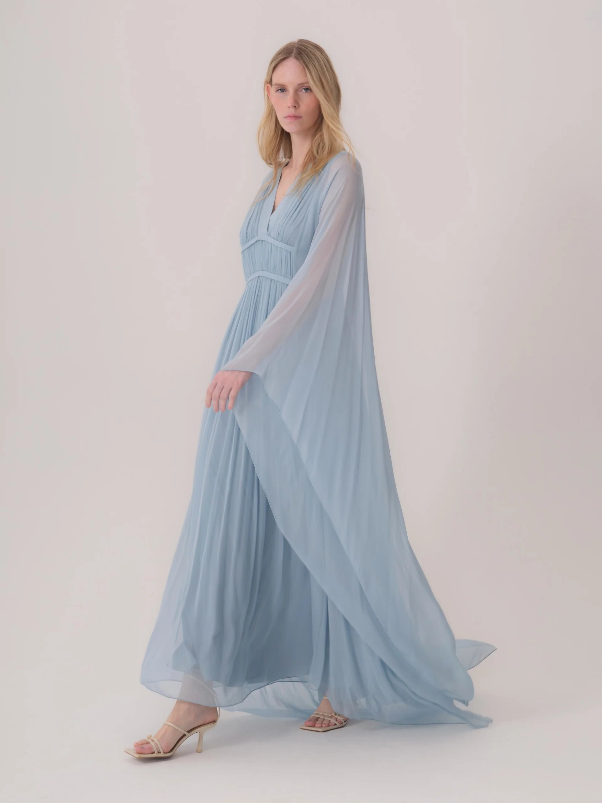 Light blue pleated maxi dress with a V-neck, empire waist, and long, flowing sheer cape-like sleeves.