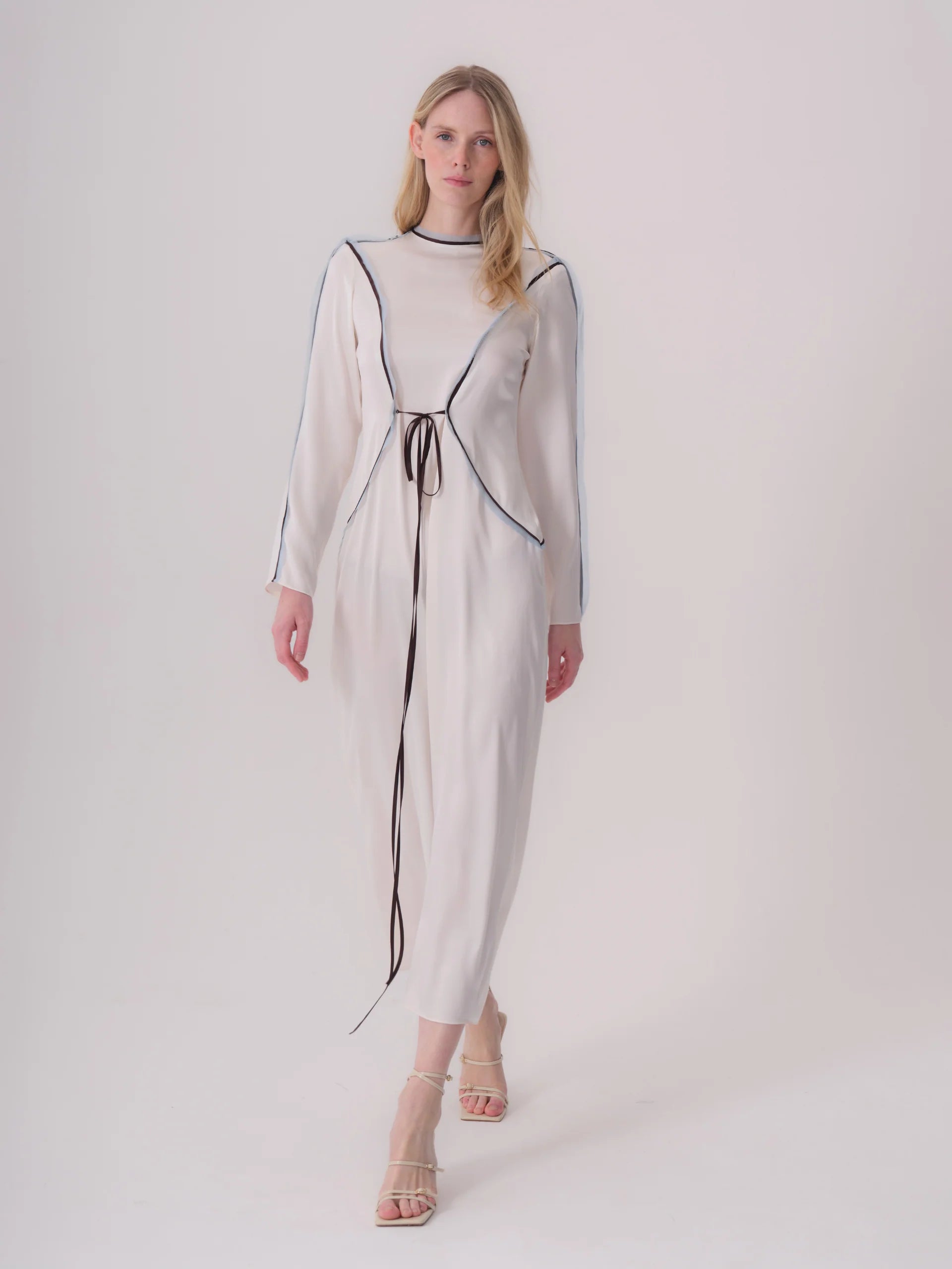 Ecru long-sleeved midi dress with black piping outlining seams on the bodice and sleeves, and a black waist tie.