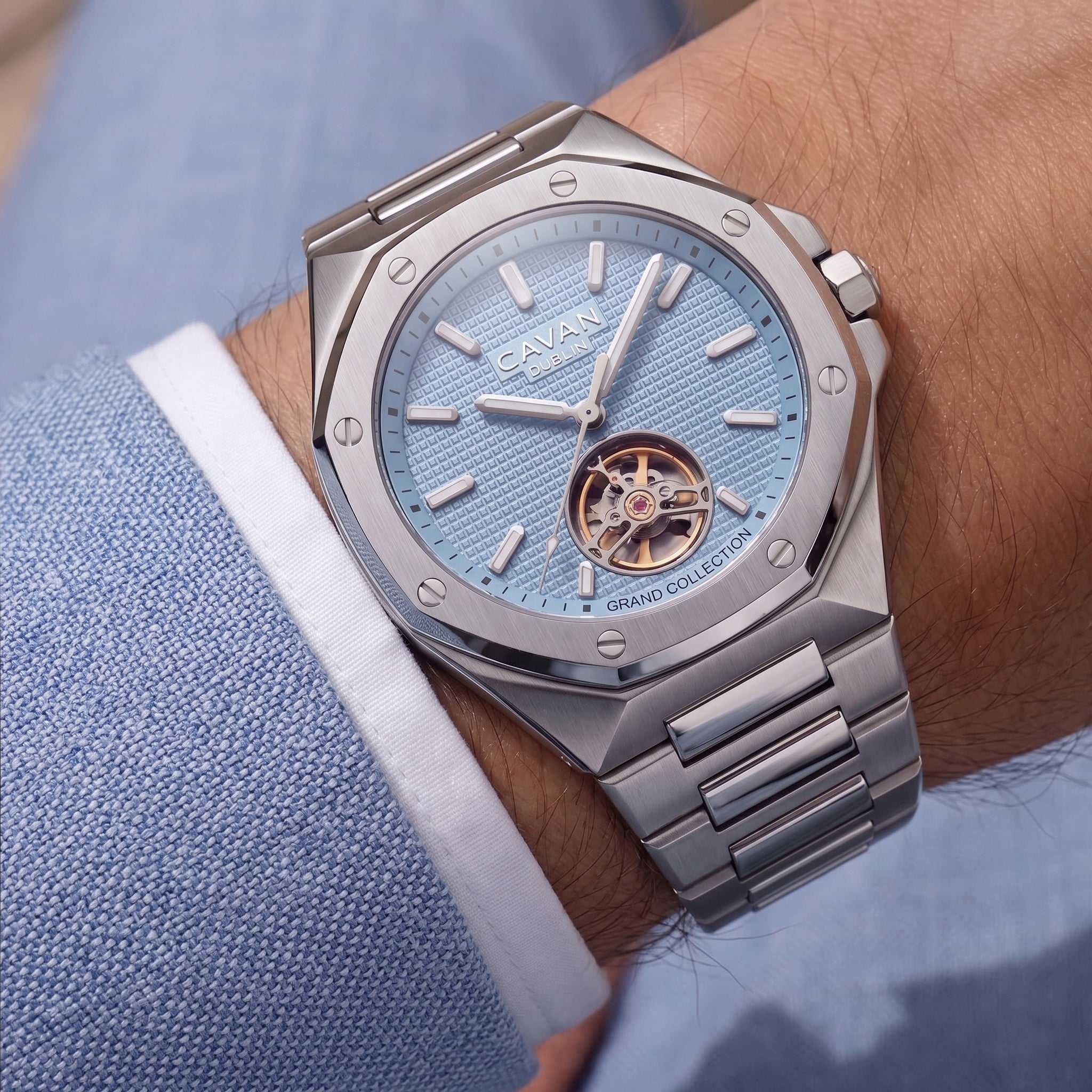 Silver-toned octagonal watch with light blue textured dial, visible gold-toned tourbillon, and silver-toned metal link bracel