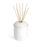 White cylindrical reed diffuser with a rounded top, gold-toned band, and light wooden reeds. Embossed logo on front.