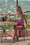 A woman sits on a metal chair on a balcony wearing a purple satin slip with green lace trim and green high-heeled sandals. A small table with flowers and bread is beside her, with a scenic outdoor background.