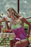 A woman wearing a purple satin slip dress with green lace trim sits on a balcony next to a small table with flowers and pastries.