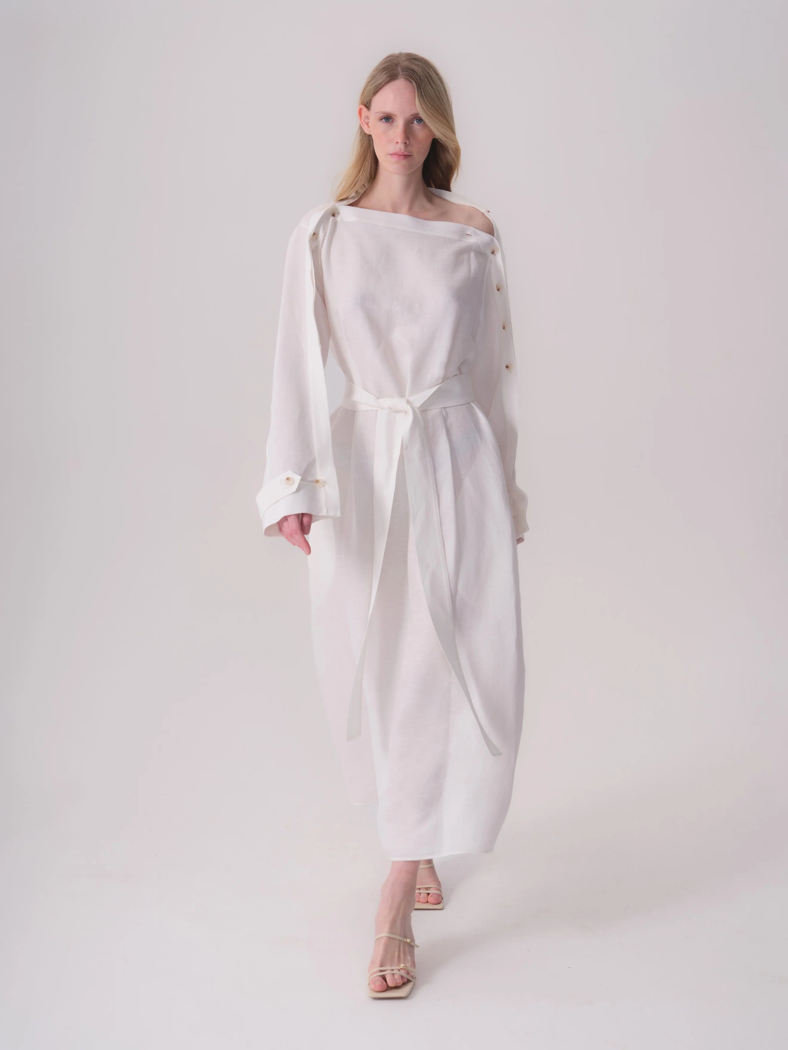 White linen-blend dress with an asymmetric neckline, long sleeves, and a matching belt. Light brown buttons on left shou