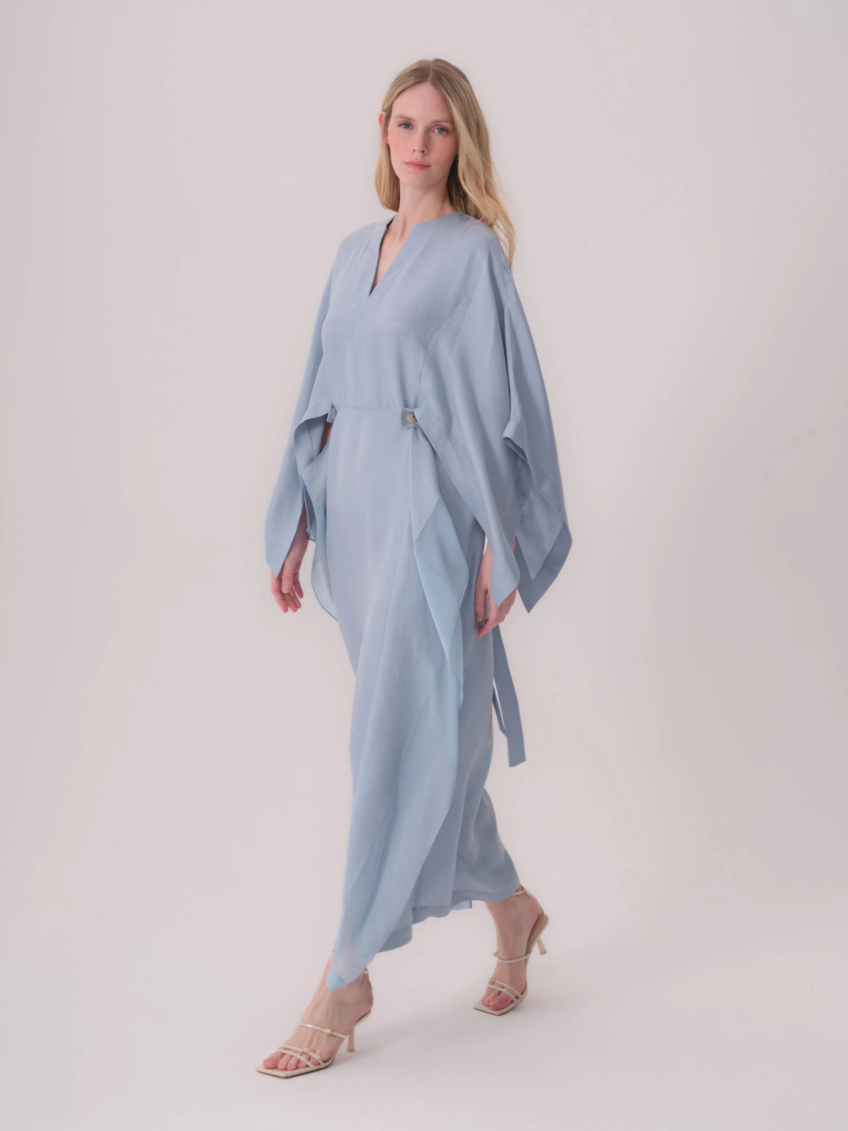 Light blue V-neck maxi dress with wide, flowing sleeves, a gold-toned button detail at the waist, and a tie belt.