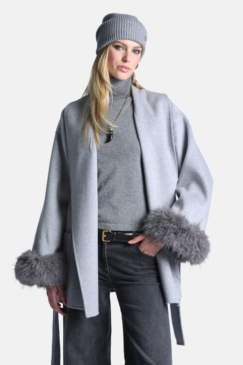 A woman modeling a light gray open-front coat with gray faux-fur trimmed cuffs, worn over a gray turtleneck, dark gray jeans, a gray beanie, and a black belt, against a plain light gray studio background.