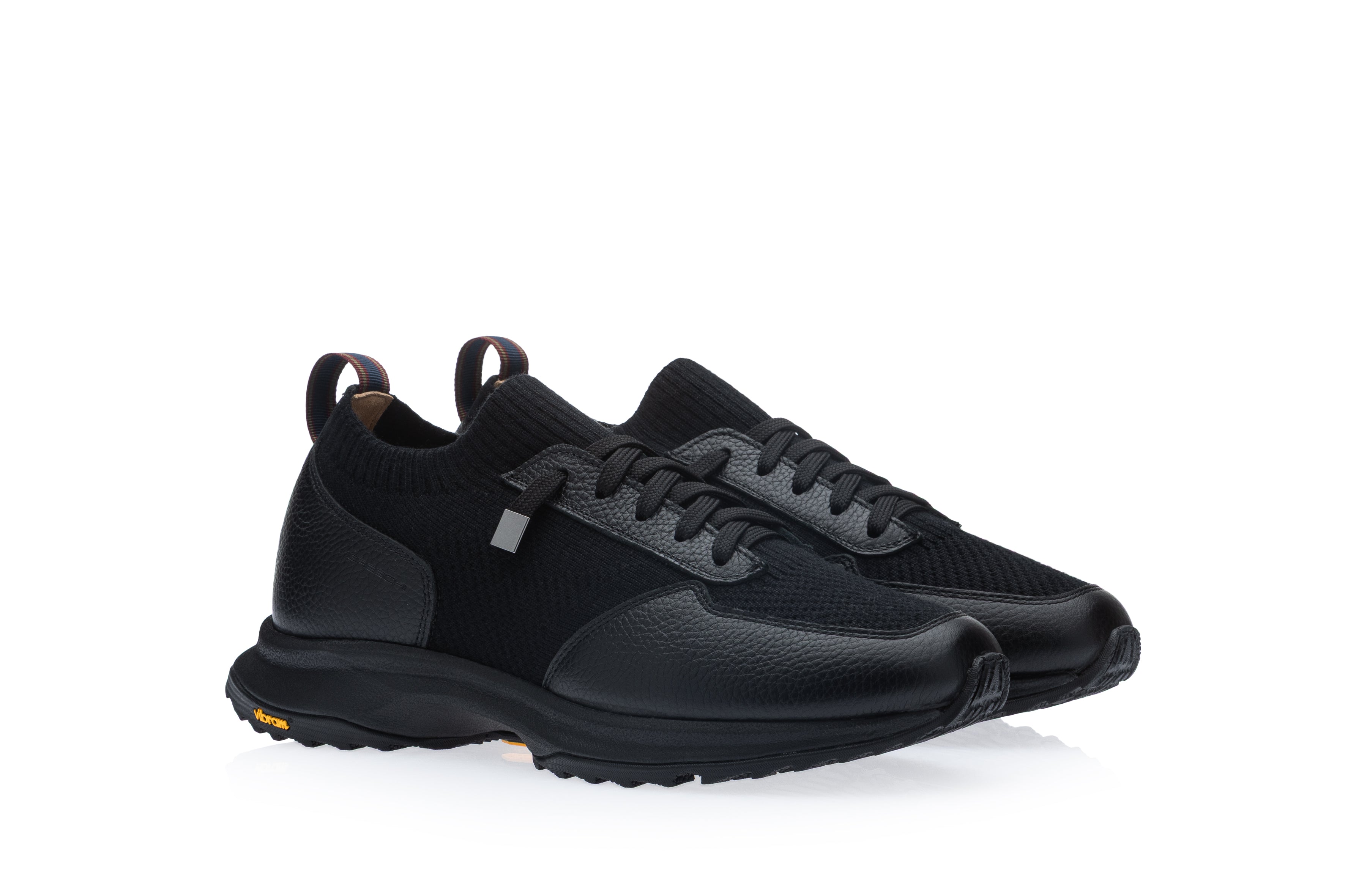 Pair of black sneakers featuring knitted fabric, pebbled leather, and mesh panels. Black laces with a silver-toned tag. Strip