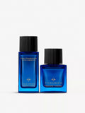 Two blue glass bottles, black caps. One tall rectangular, labeled 'Hair Fragrance with Keratin'. Shorter square. Gold-toned e