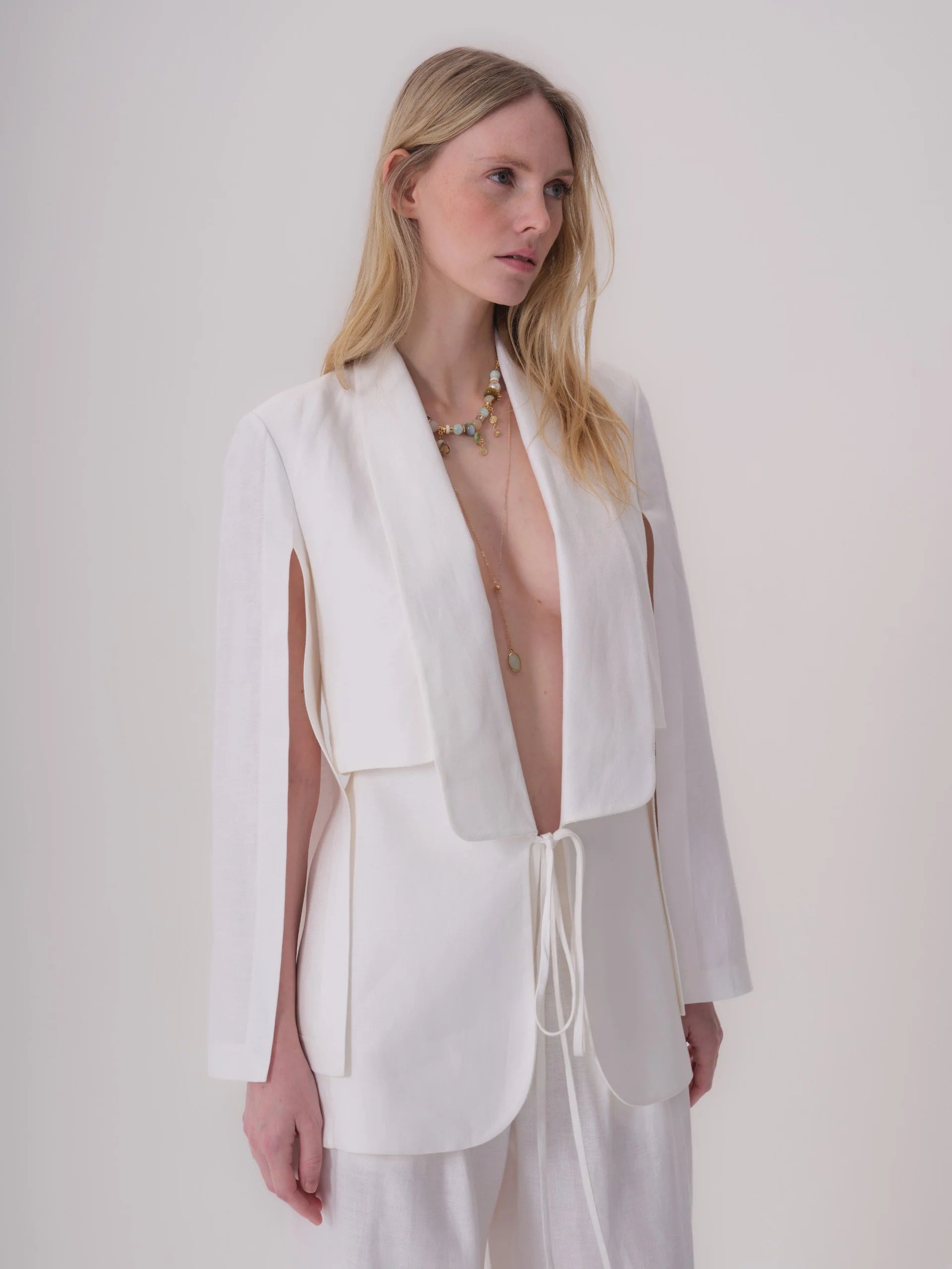 White linen-blend jacket with a deep V-neck shawl collar, layered top, long sleeves with outer arm slits, tie-front, and roun