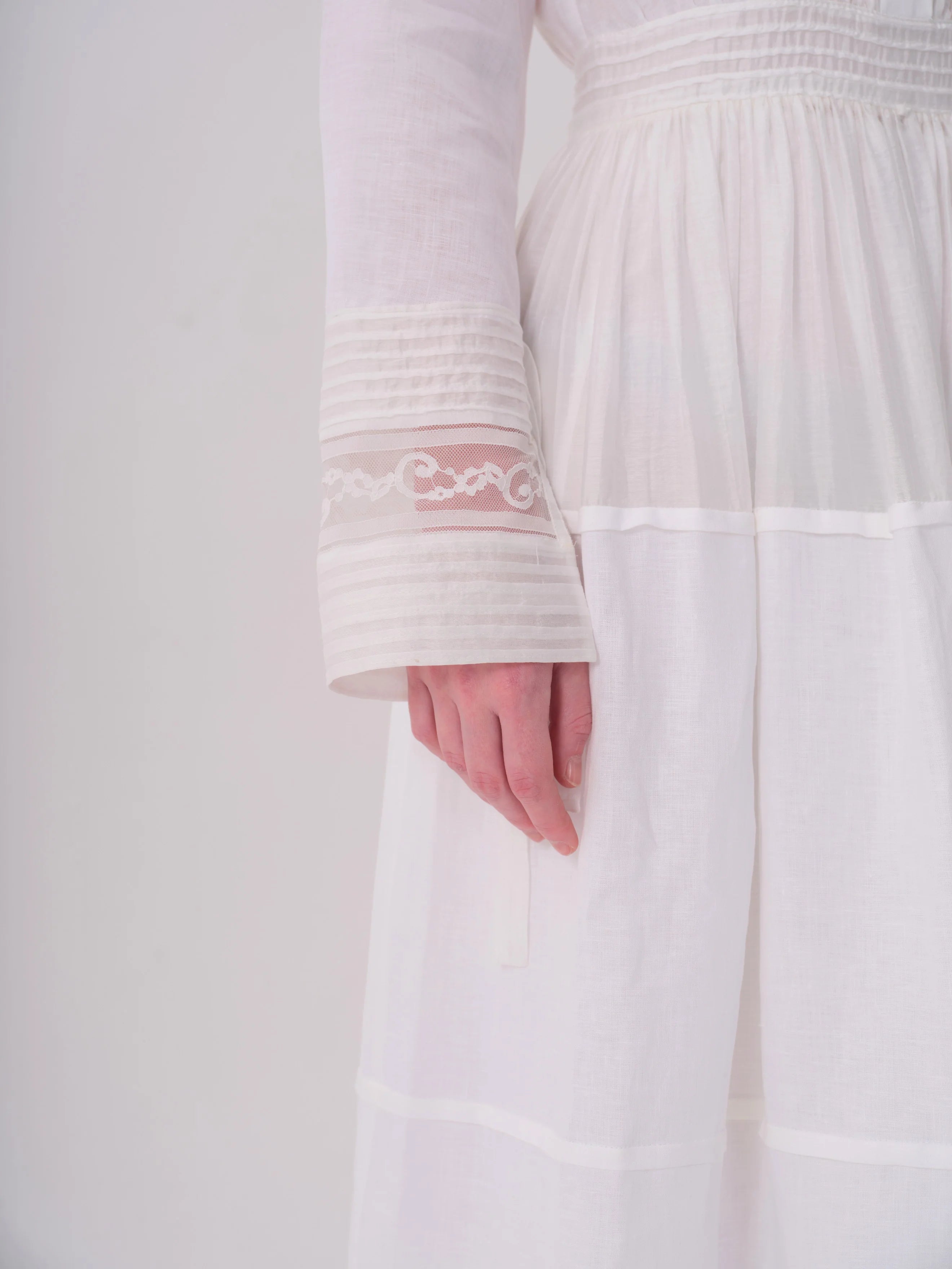 White long-sleeved dress with smocked bodice, tiered skirt. Cuffs have horizontal pintucks and a sheer lace scroll panel.