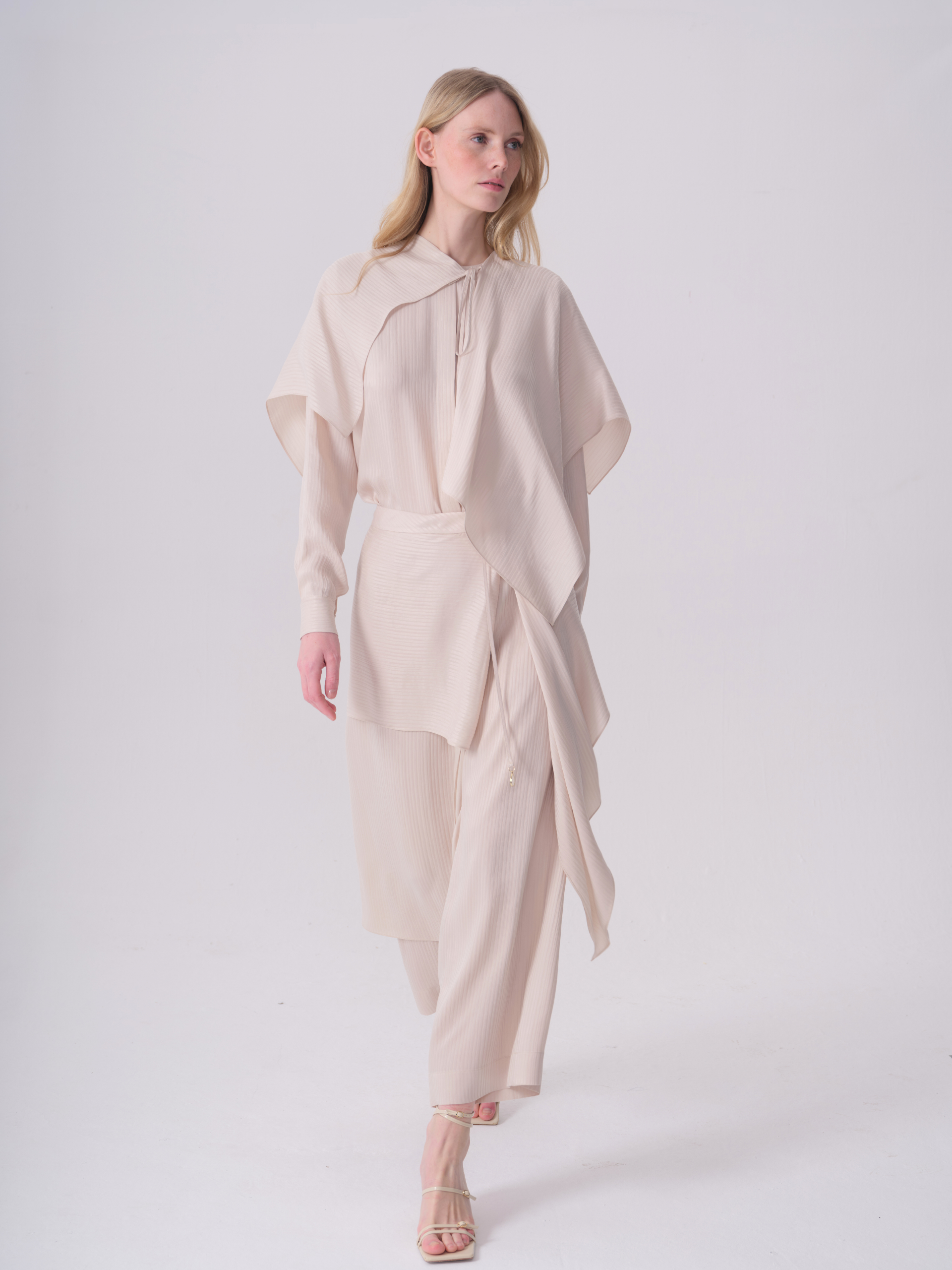 Off-white ribbed straight-leg trousers, ankle-length. Features a draped panel on the left hip with a small gold-toned bead.