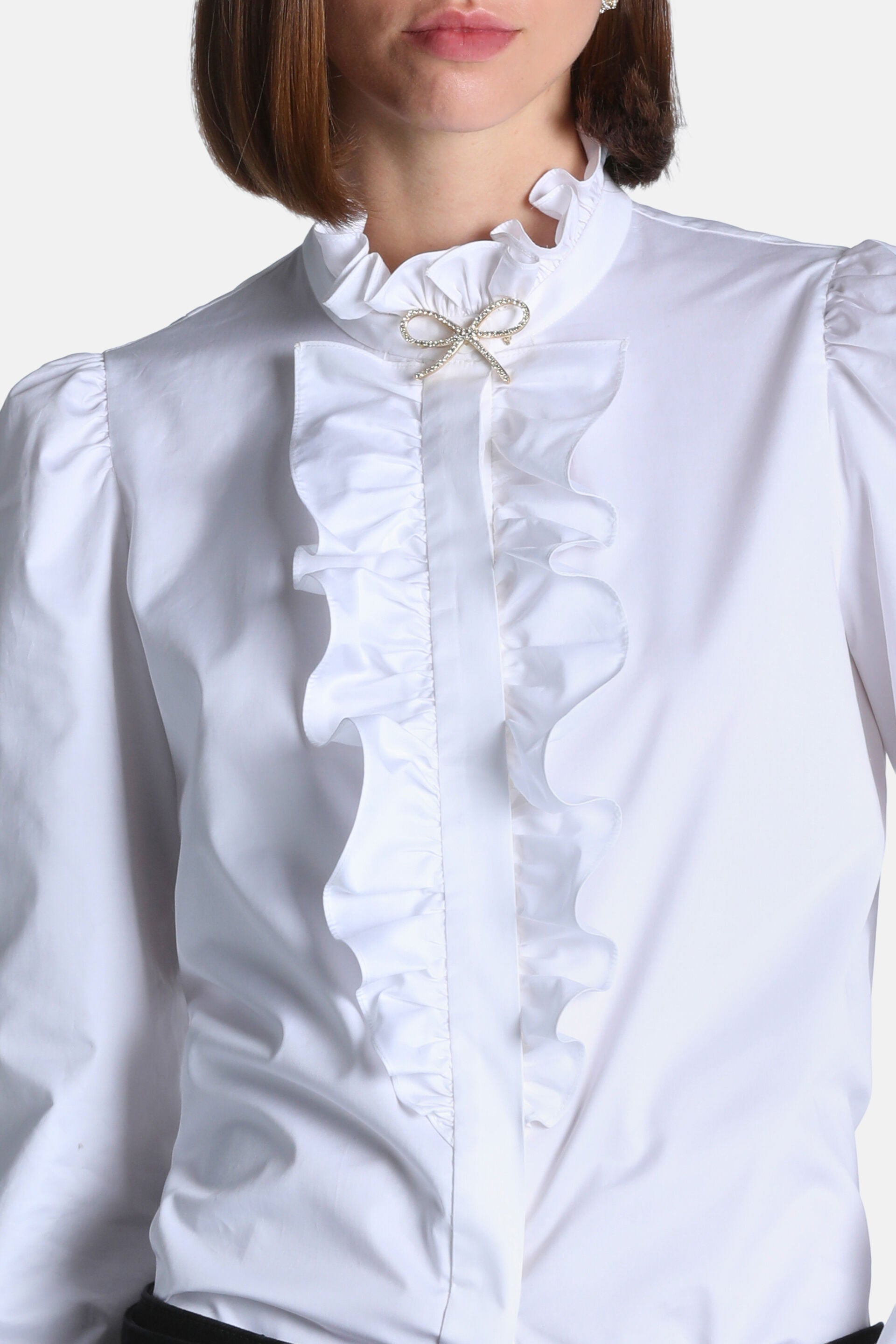 White shirt with a ruffled stand-up collar, ruffled front placket, and a gold-toned crystal bow brooch. Puffed shoulders.
