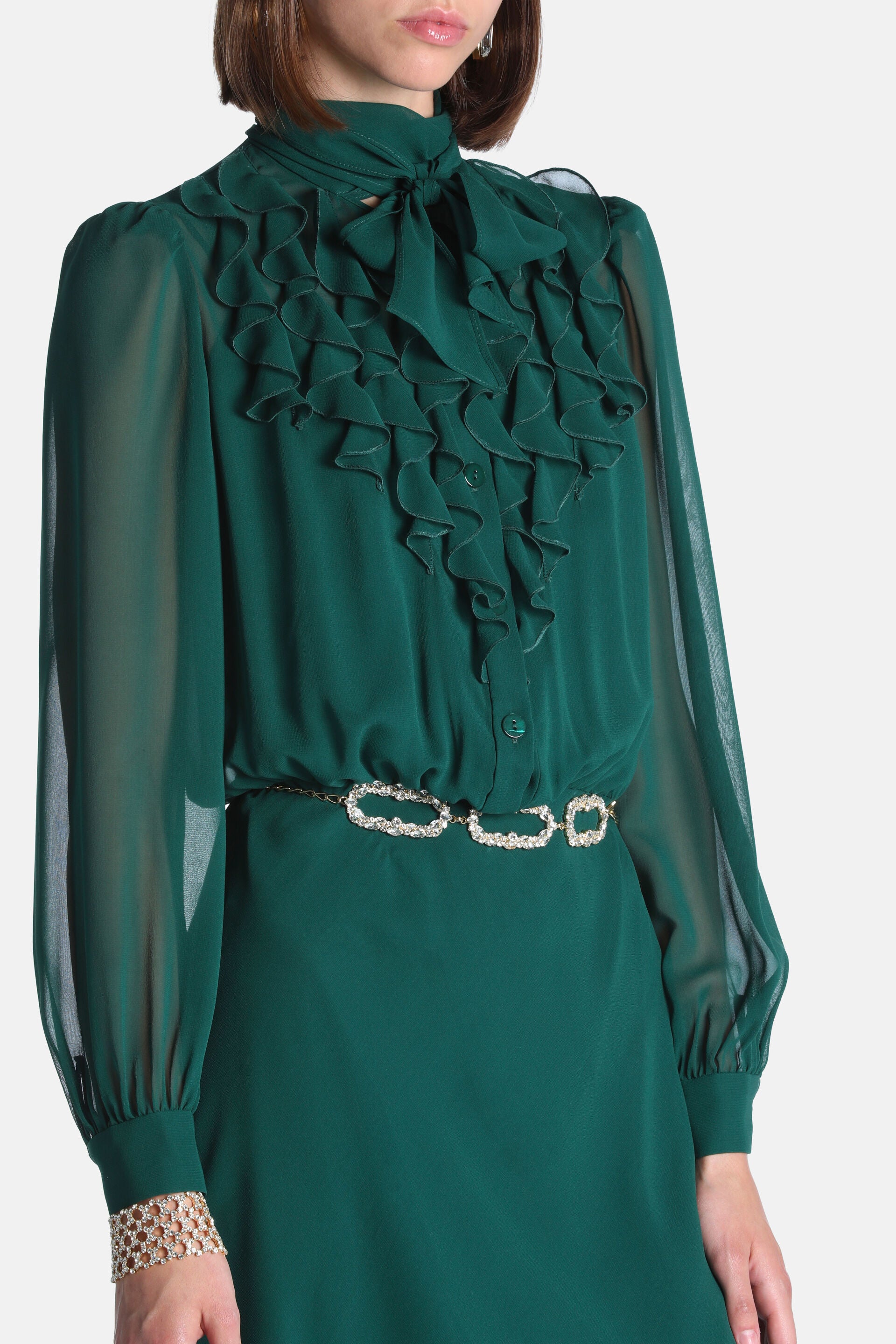 Dark green dress with a high tie-neck, ruffled bodice, sheer long sleeves, and a gold-toned crystal-embellished chain belt.