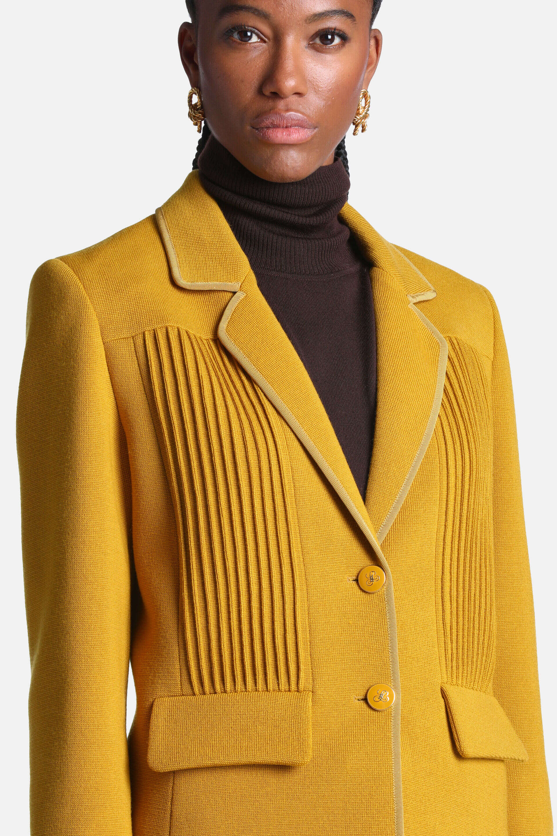 Mustard yellow knit jacket with notched lapels, lighter trim, vertical pleats on the chest, two front buttons, and flap pocke