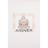 White fabric with a beige cartoon bear wearing a patterned hat with a gold-toned A-logo, framed by a beige A-logo border. Bla