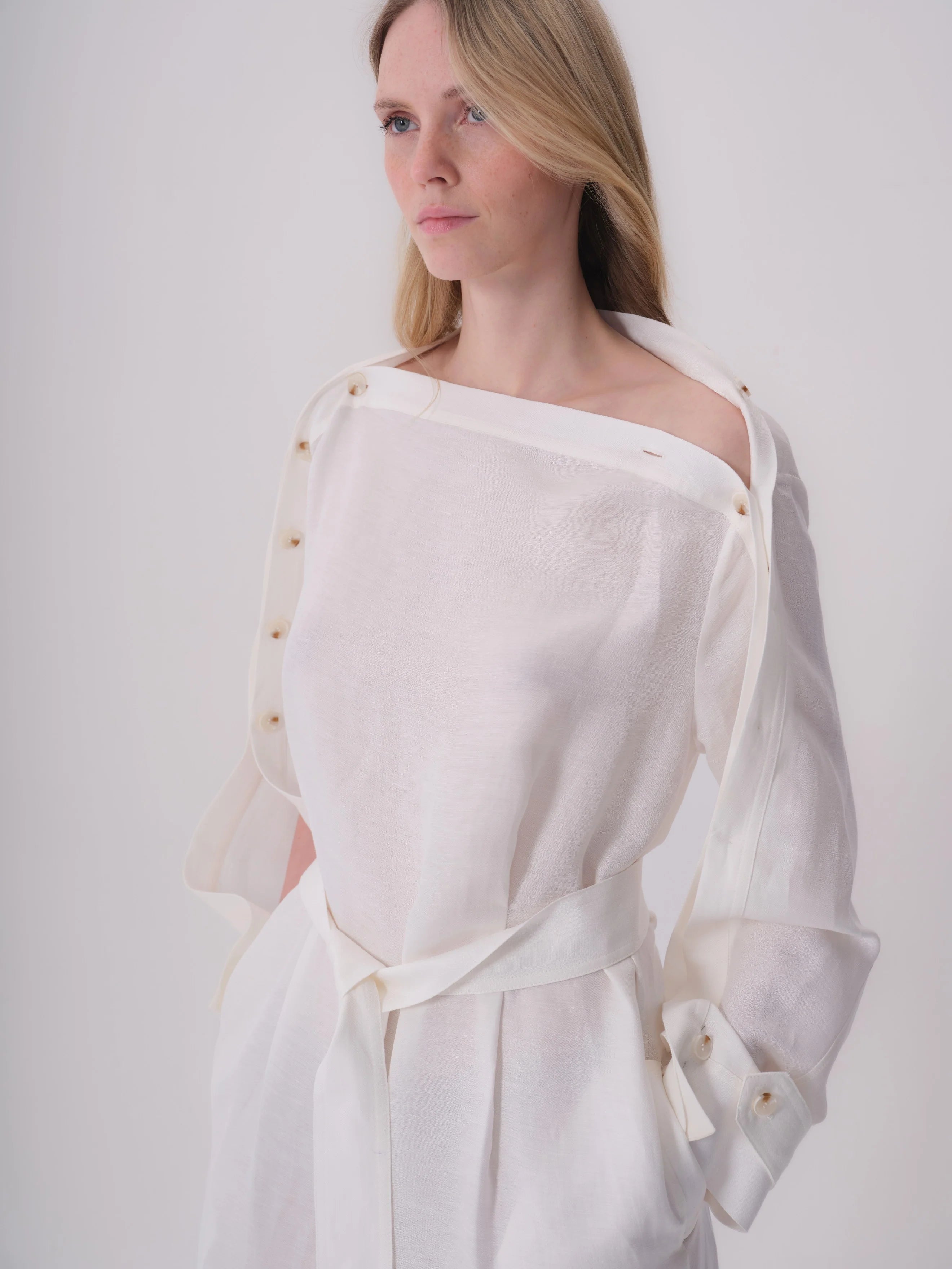 White linen dress, asymmetrical off-shoulder, long sleeves with rolled buttoned cuffs, tied waist belt, light brown buttons.