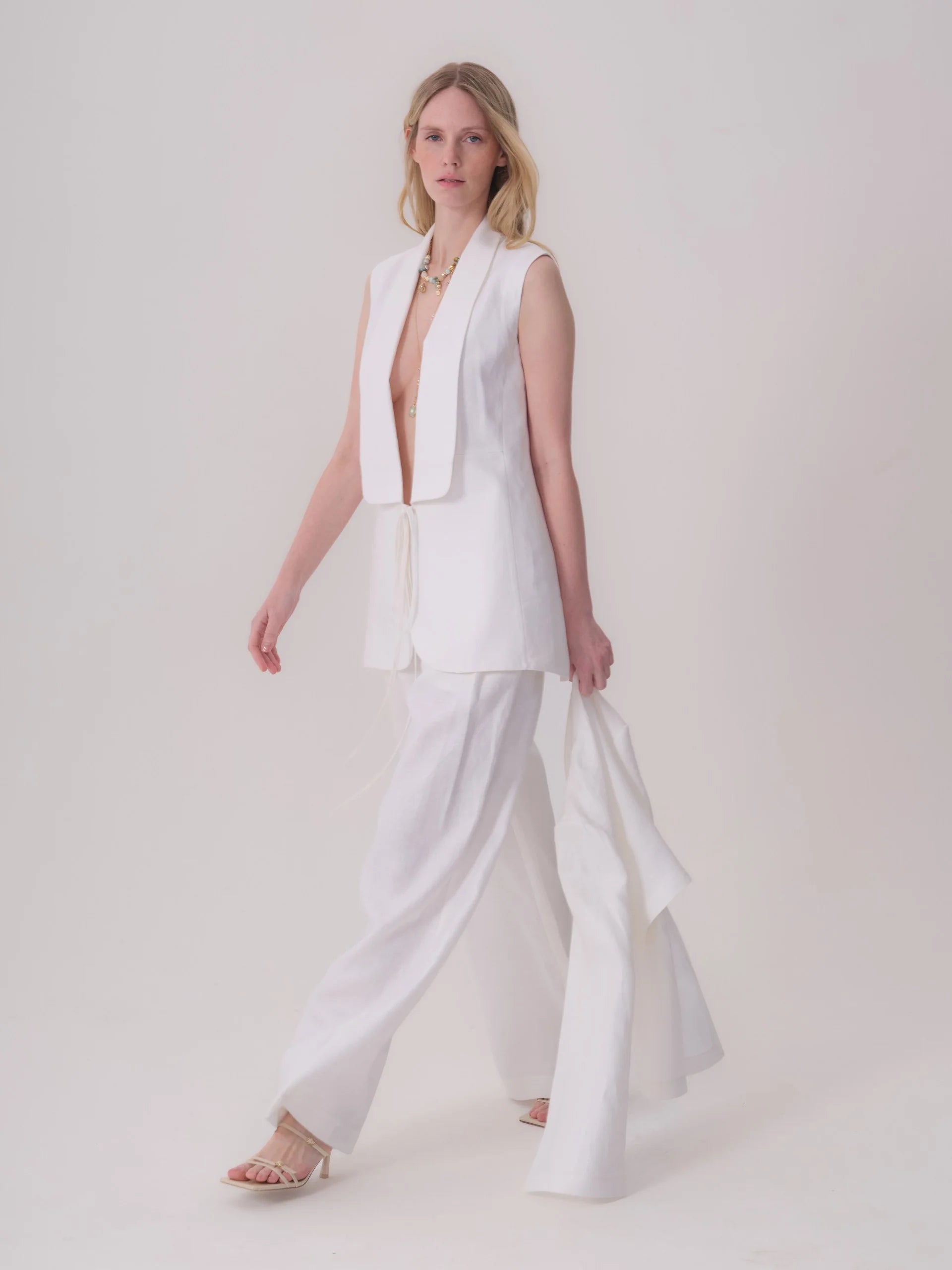 White sleeveless jacket with wide lapels, deep V-neck, and front tie closure. Made of textured fabric.
