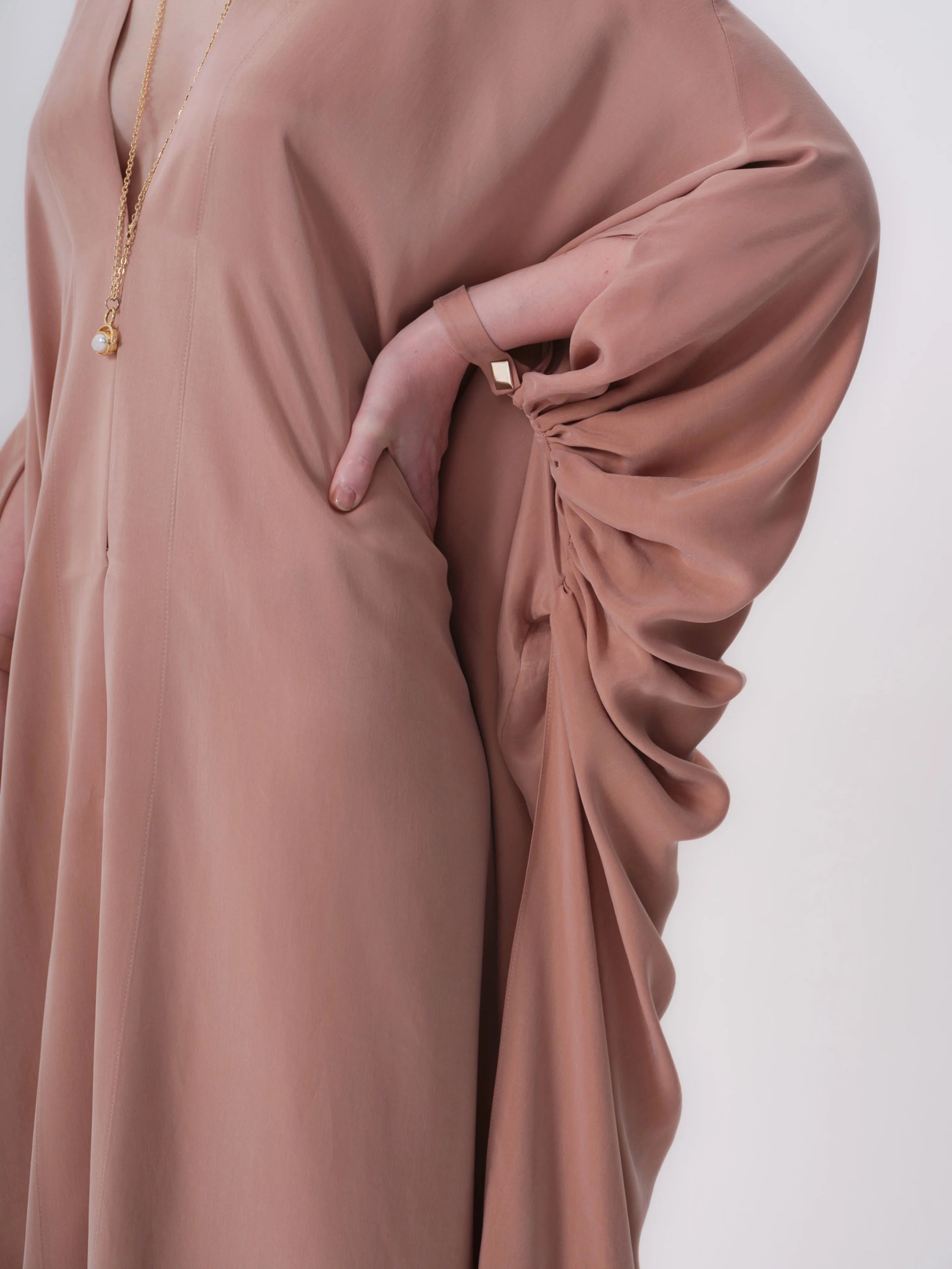 Dark beige abaya with V-neck. Loose fit, long sleeves; right sleeve has ruched draping. Vertical front seam and side pocket.