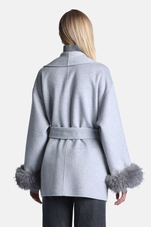 A woman shown from the back wearing a light gray belted coat with gray faux-fur trimmed cuffs, paired with dark gray pants, against a plain light gray background.